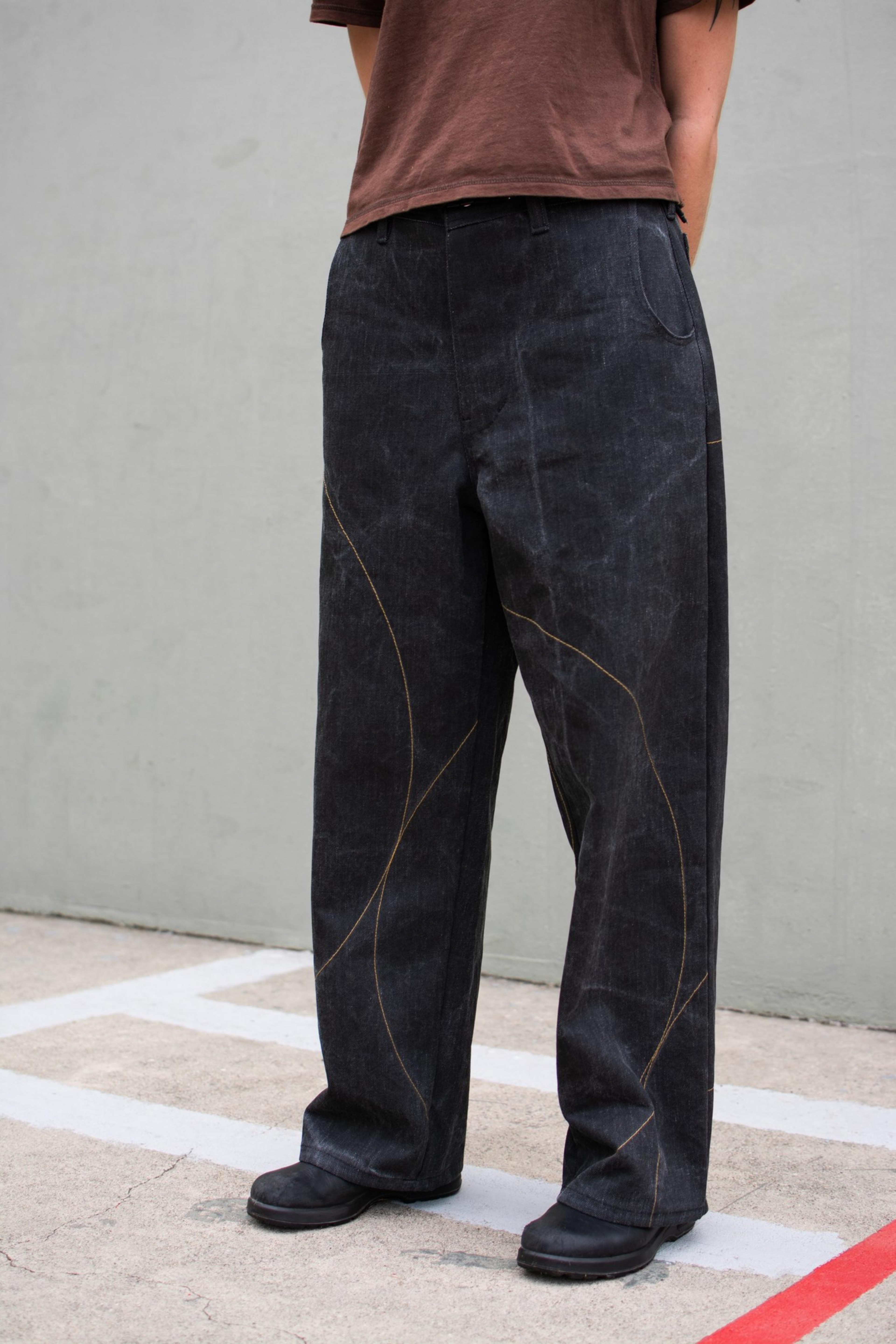 A person wears loose black pants with curved tan stitching and black shoes, paired with a brown shirt, standing on concrete with white and red lines.