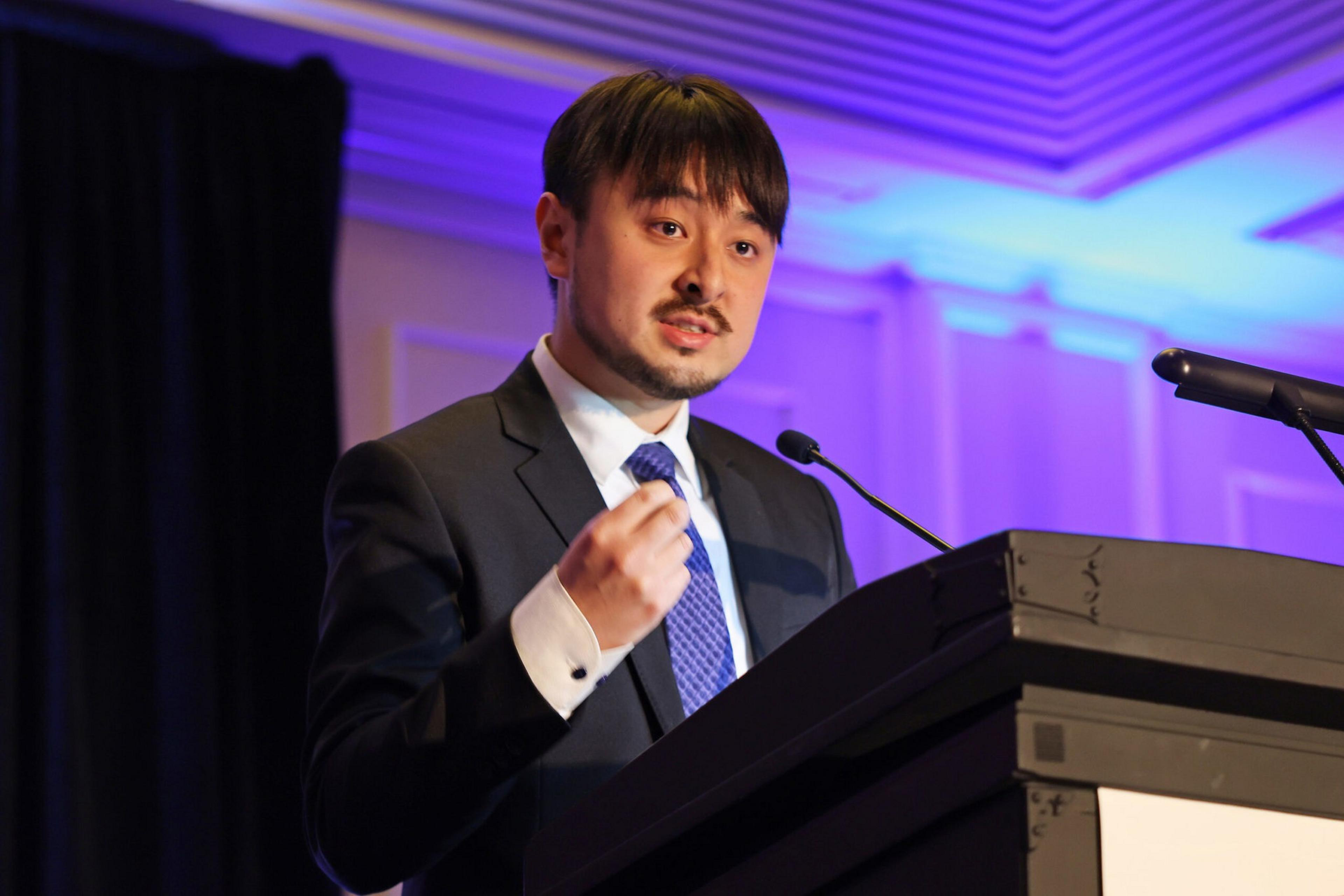 A man in a suit and blue tie is speaking and gesturing at a podium with a microphone in a room lit by blue and purple lights.