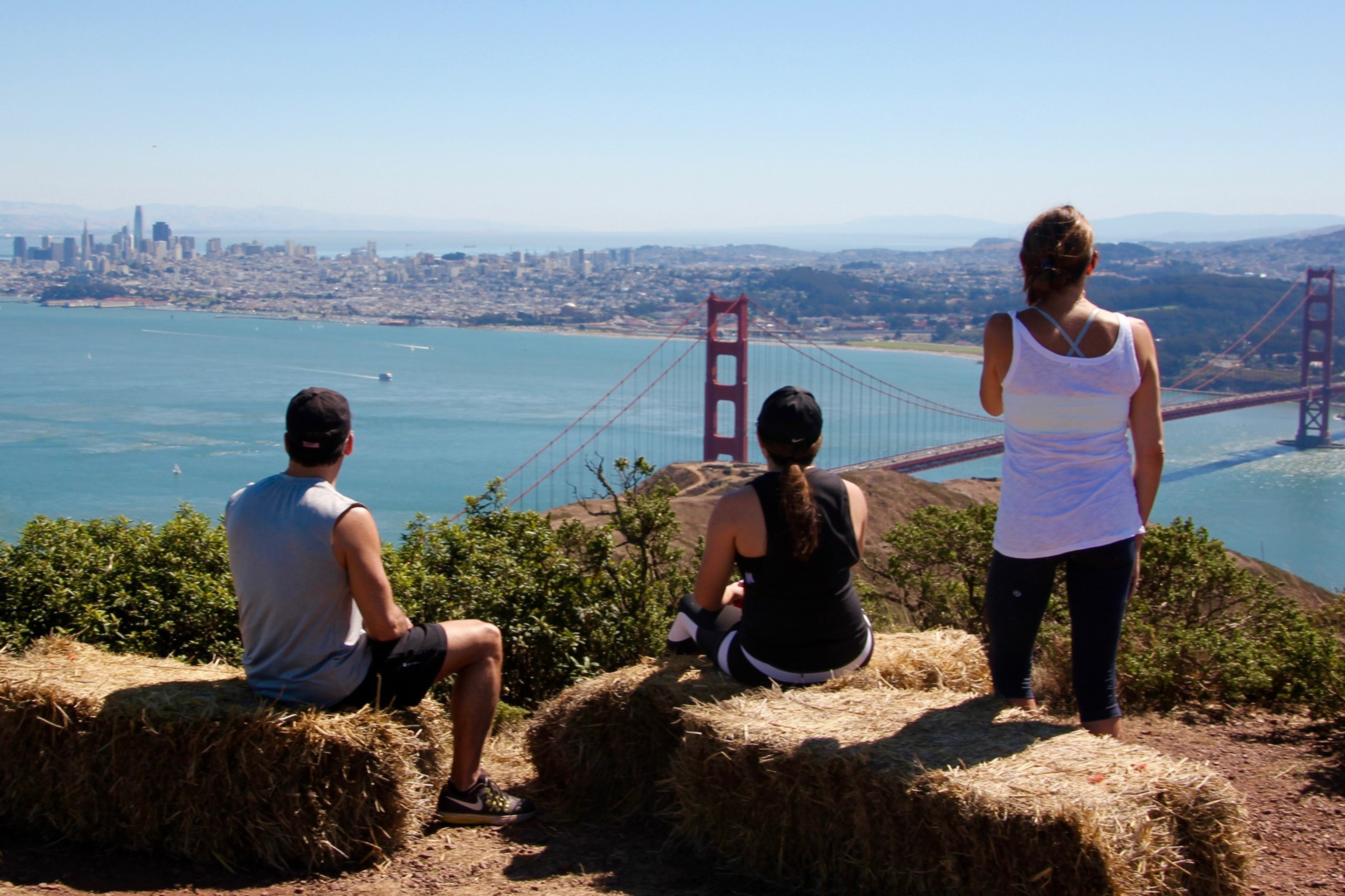 Three people overlook the Golden Gate Bridge and San Francisco skyline from a hill with hay bales.