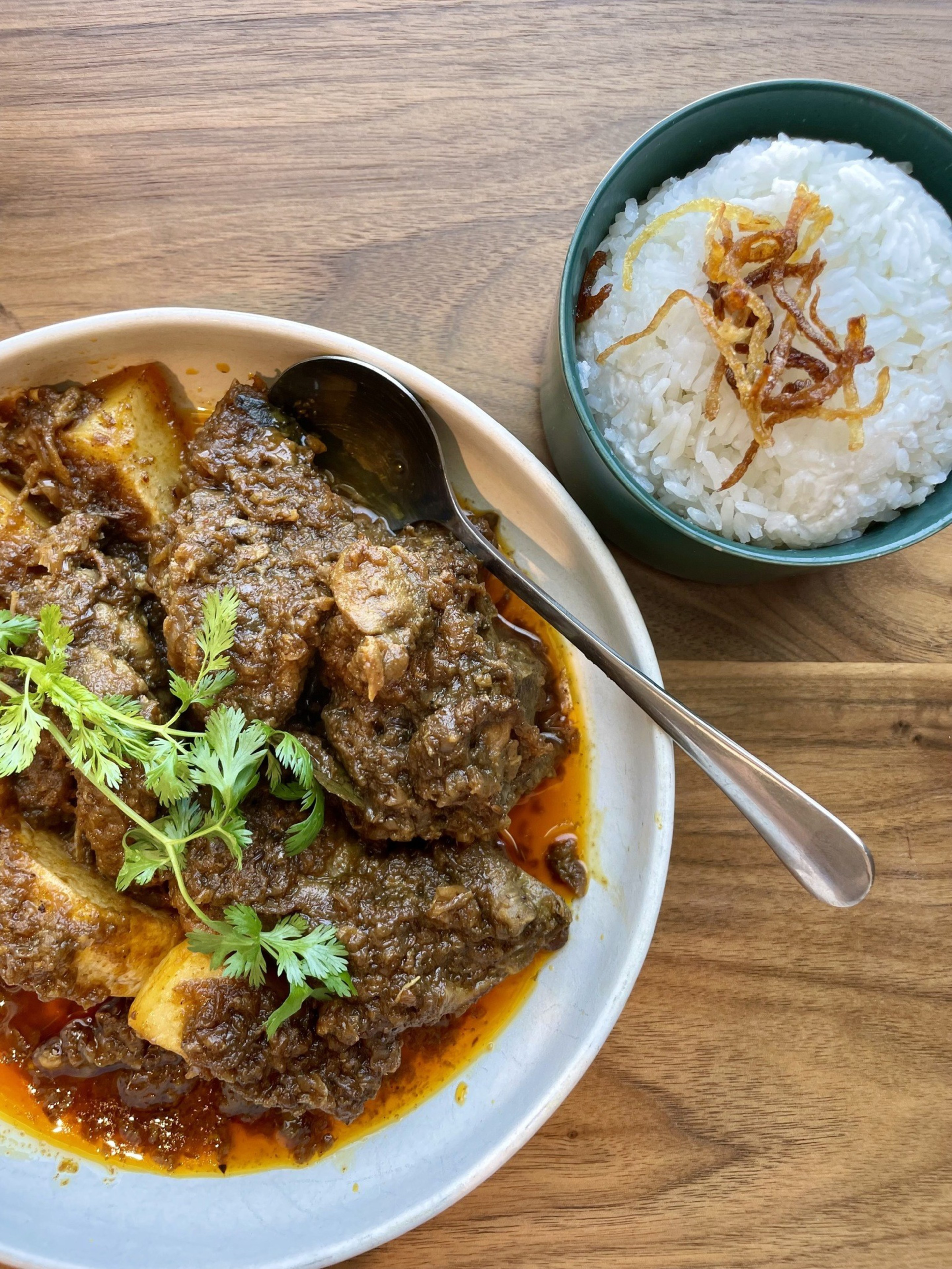 A white bowl of chicken curry garnished with cilantro next to a bowl of rice on a wooden table.