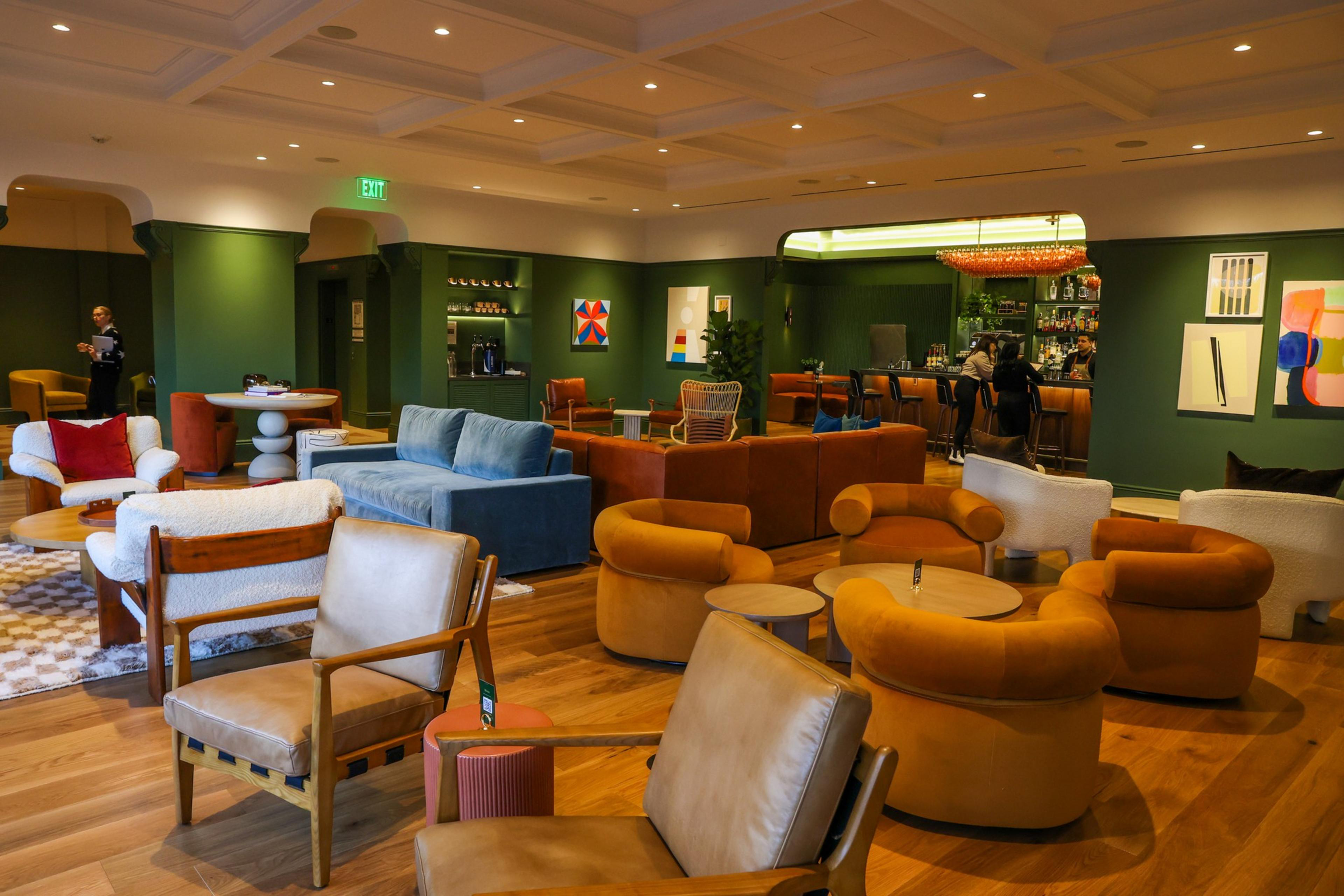 A cozy lounge with green walls features a mix of orange, blue, and white sofas and chairs around wooden tables, with a bar and abstract art in the background.