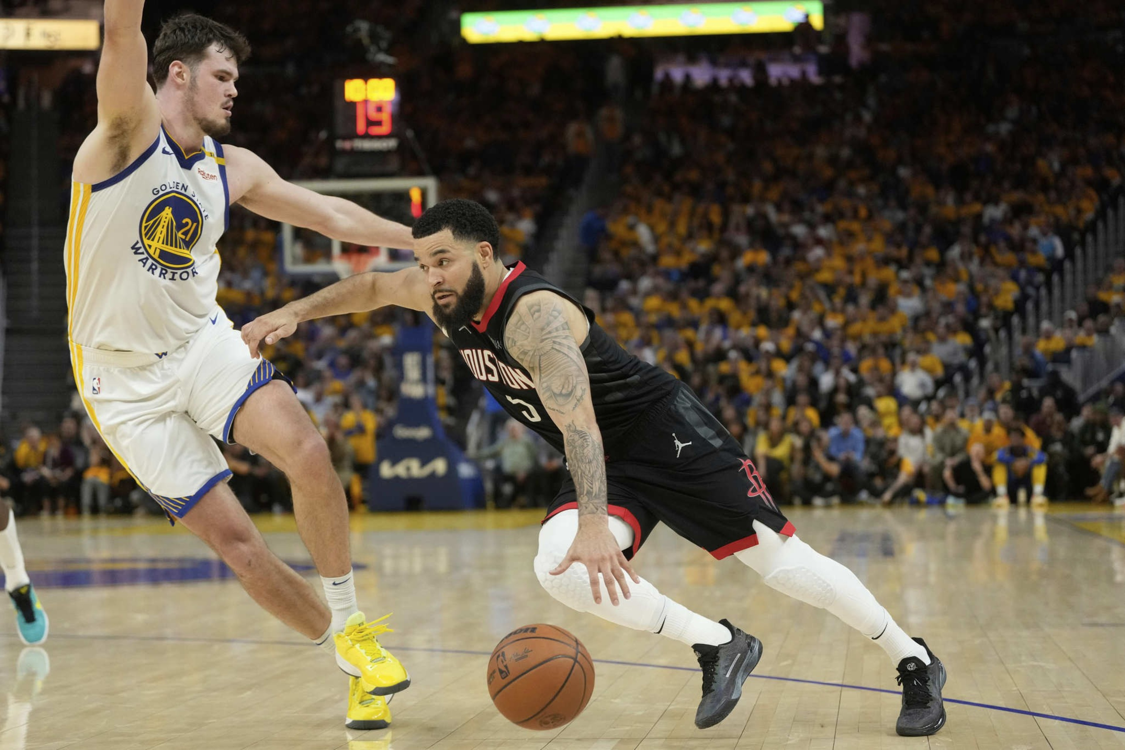 A basketball player in a black uniform dribbles past a defending player in a white and yellow uniform on a court, with a crowd in the background.