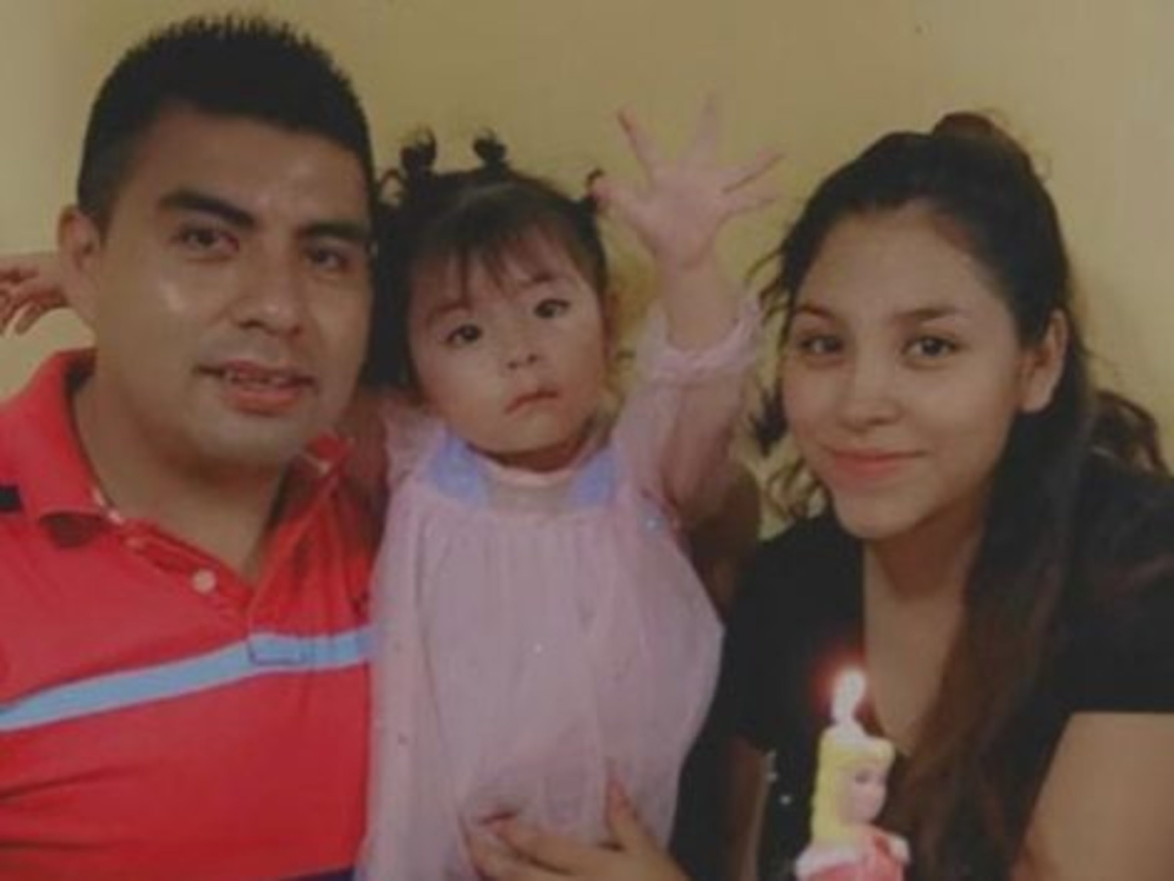 A family photo with a man, toddler, and woman; the child, raising her hand, holds a unicorn toy.