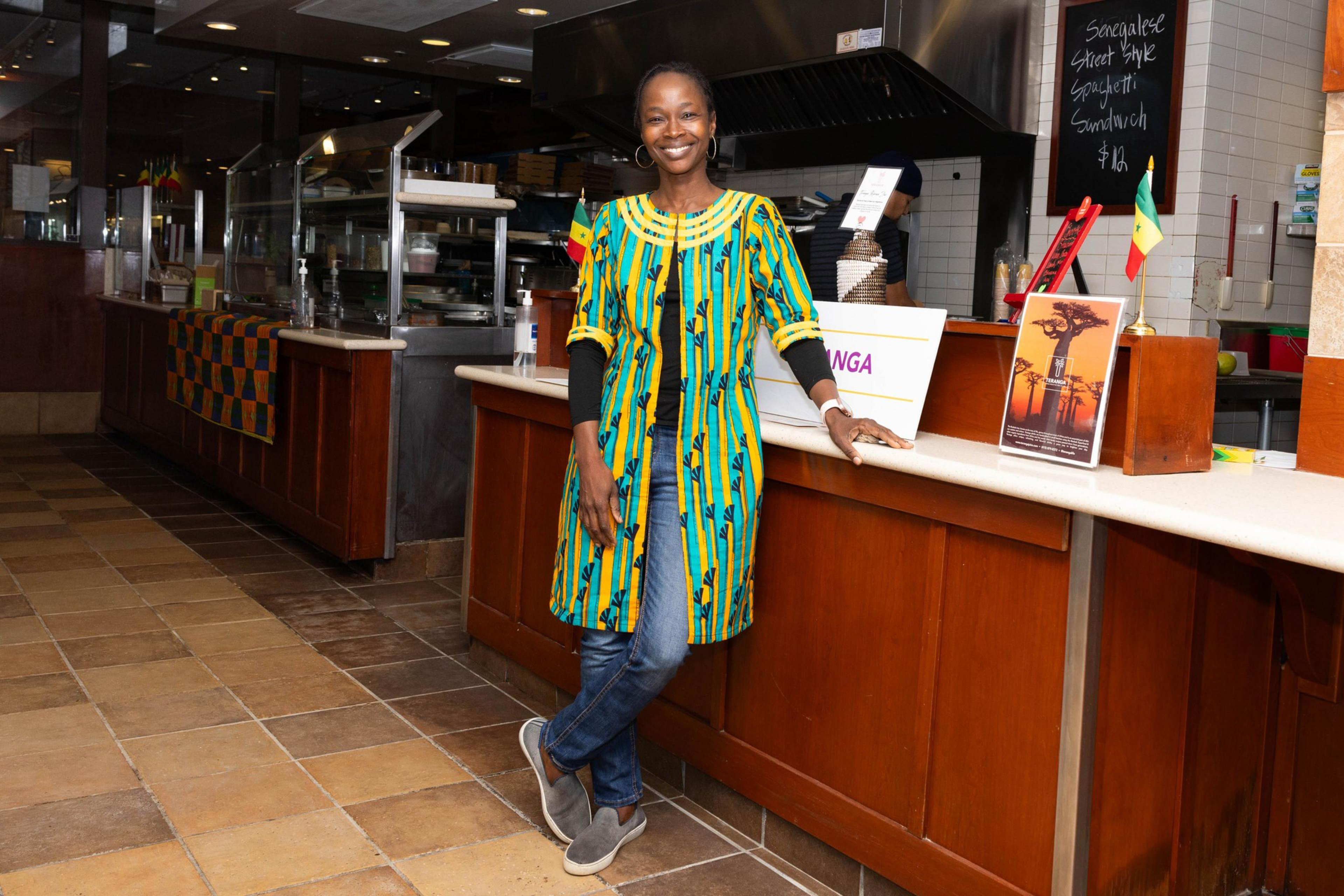 A women named Nafy Flatley, owner of Teranga, stands in the front of a counter with a kitchen setting in the background.