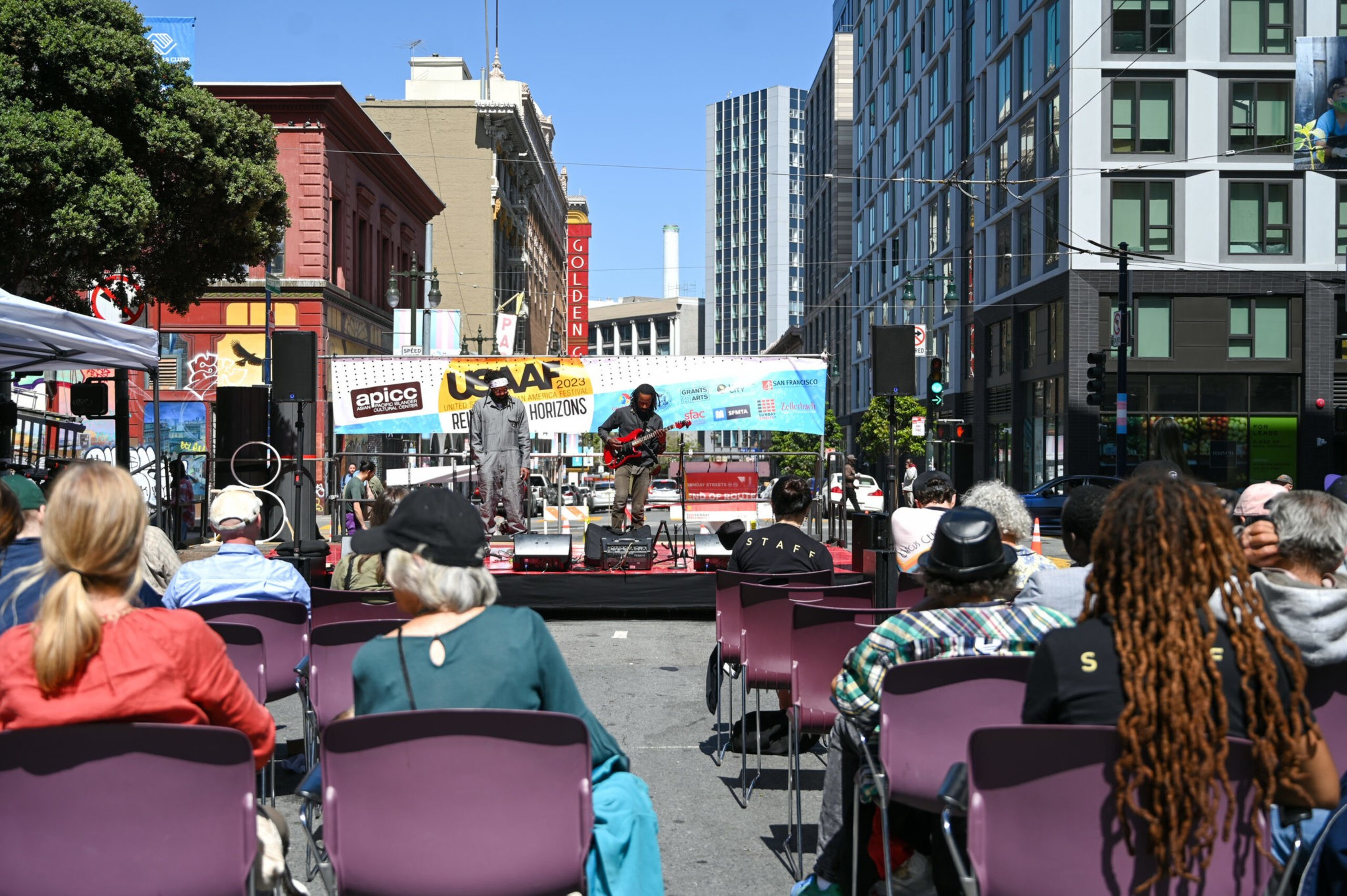 A street event with a live band on stage and onlookers seated in rows.