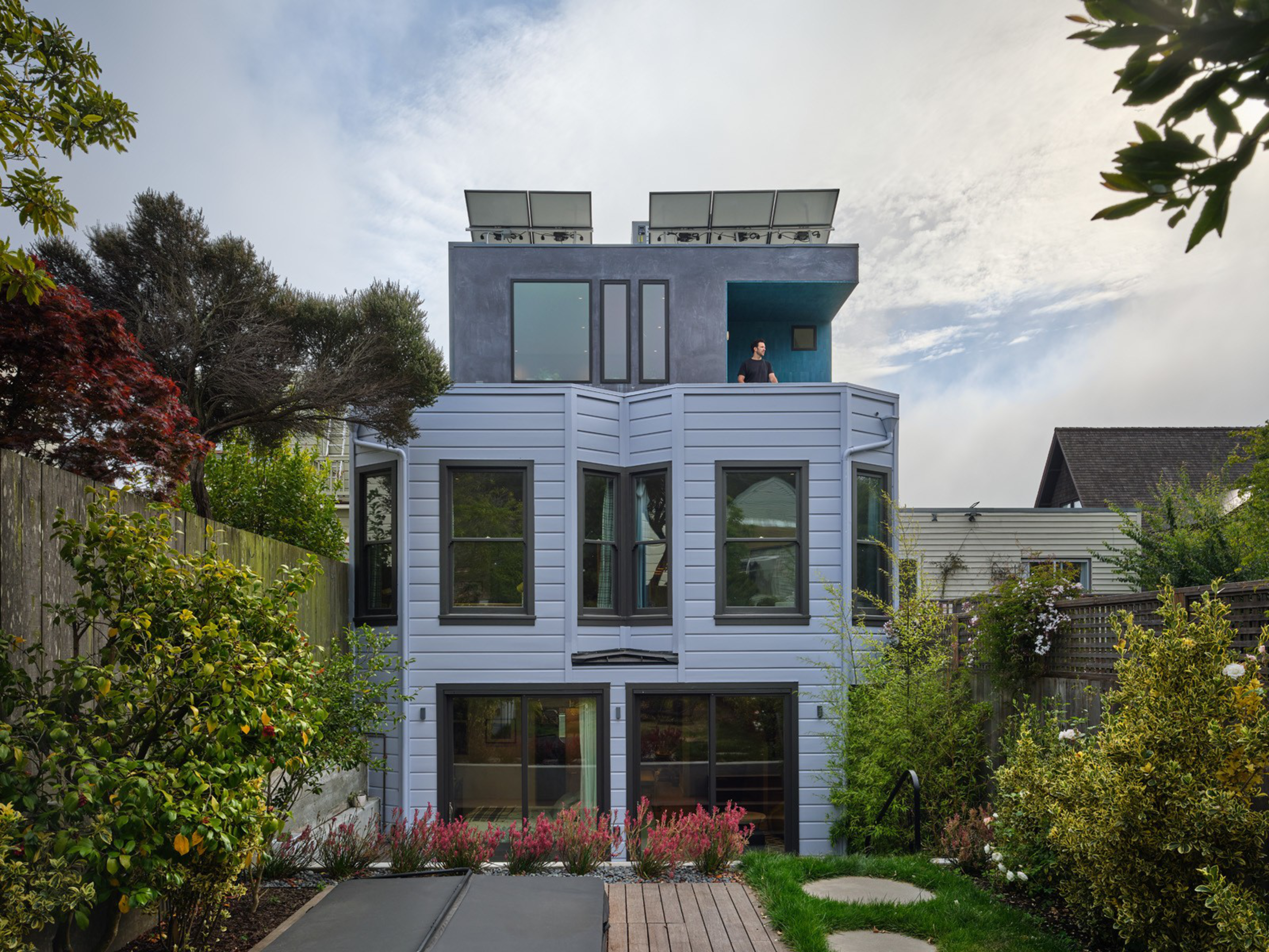 A modern three-story house with large windows, a rooftop balcony with a person standing on it, surrounded by trees and a garden pathway.