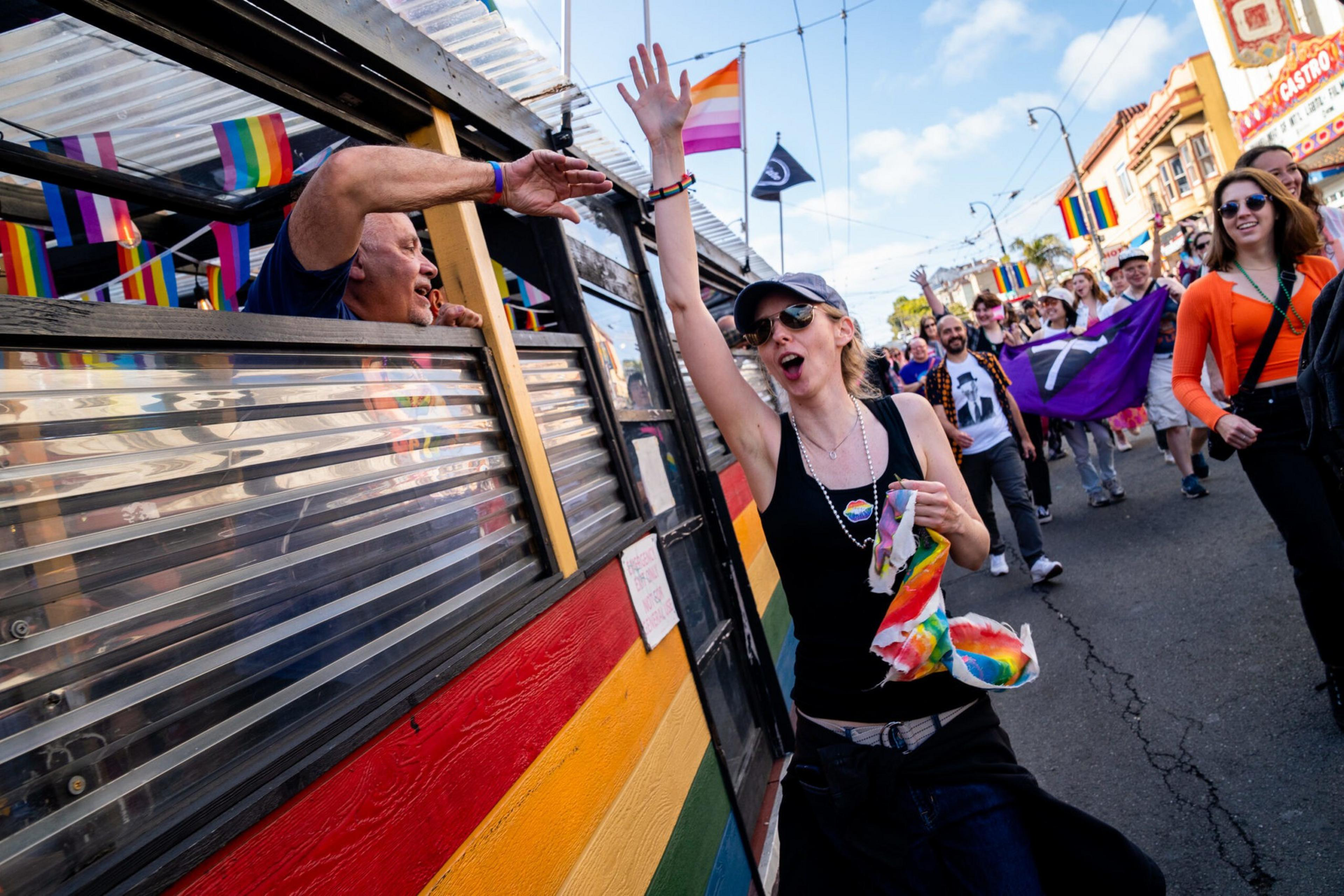 A crowd of people, some holding rainbow flags, celebrate and high-five near a decorated vehicle with rainbow colors and pride flags.