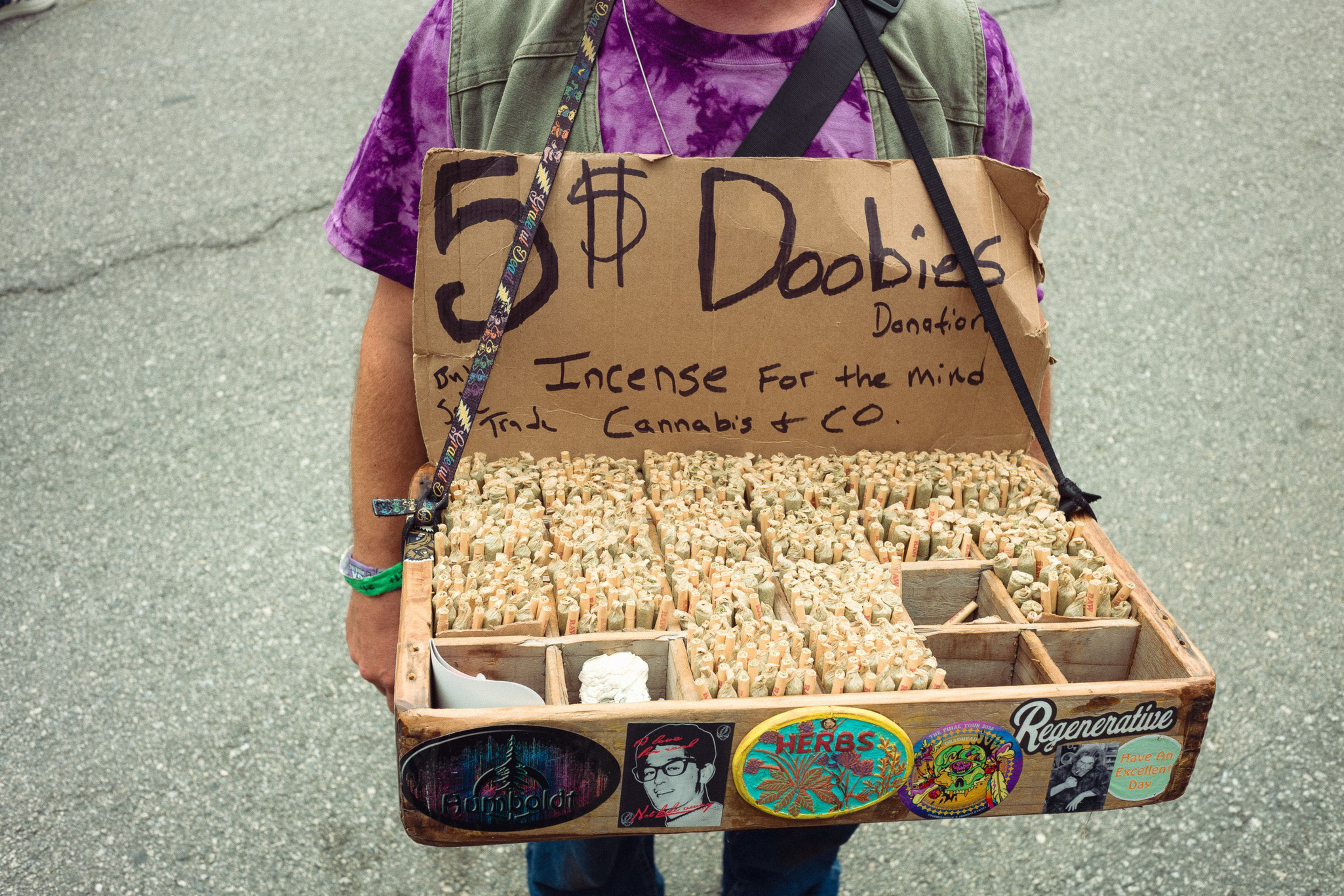 A person holds a tray filled with many hand-rolled items labeled "5$ Doobies" on a handwritten sign stating "Incense for the mind, Cannabis & Co."