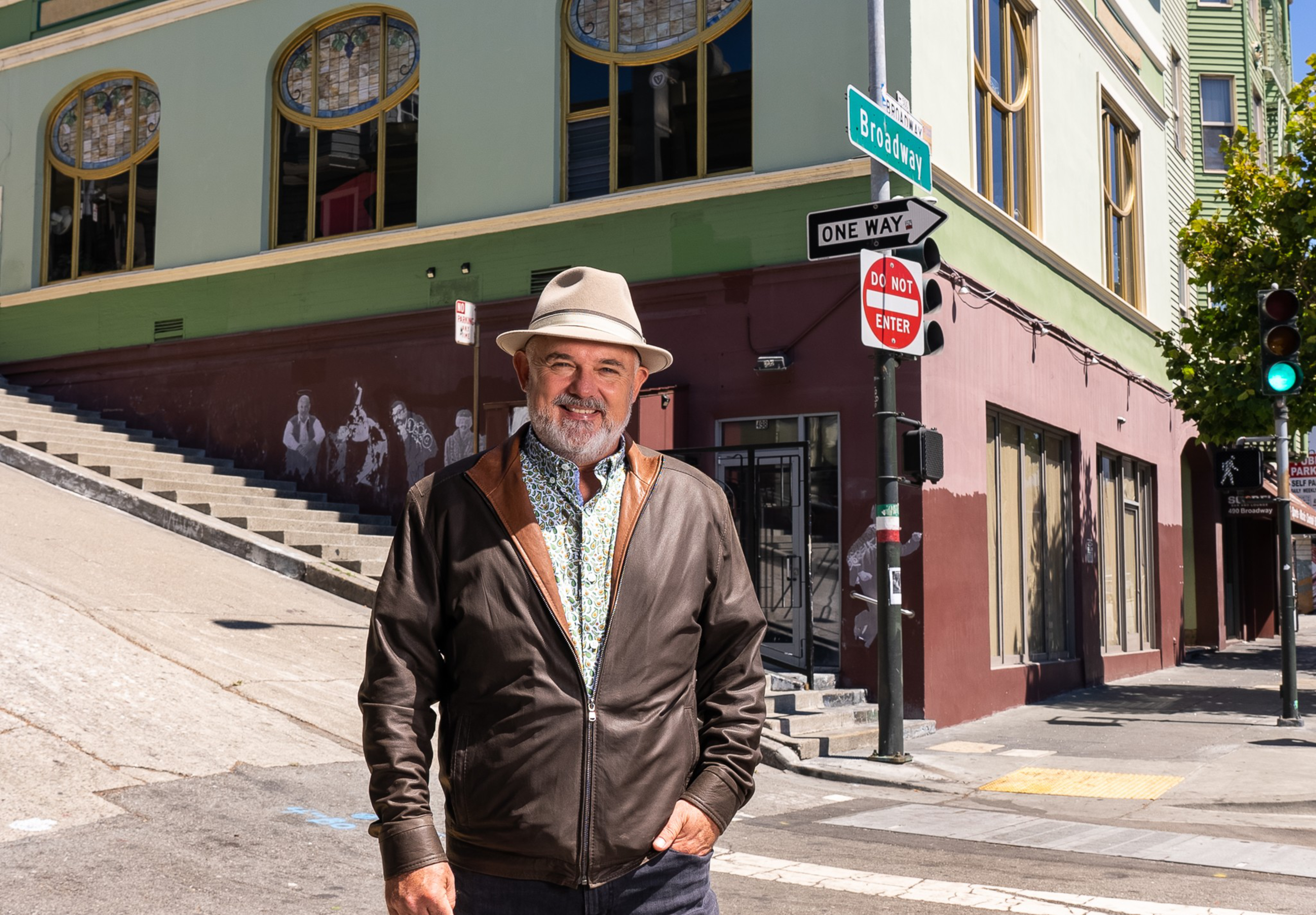 A smiling man wearing a hat and brown leather jacket stands on a sunny city street corner with a mural and street signs behind him.