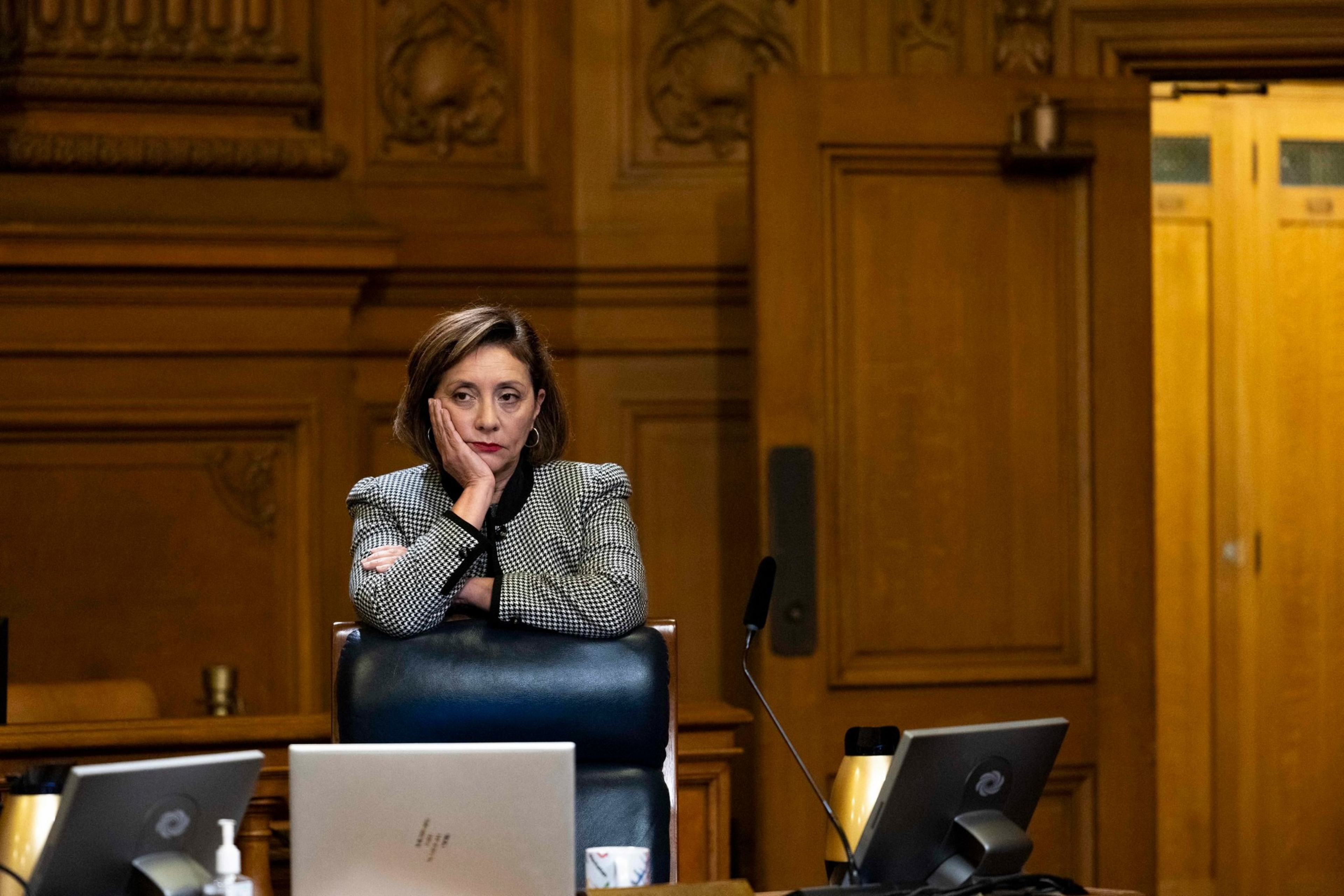 A woman in a checkered jacket leans on a chair with a hand on her face, looking thoughtful, surrounded by laptops in an ornate wood-paneled room.