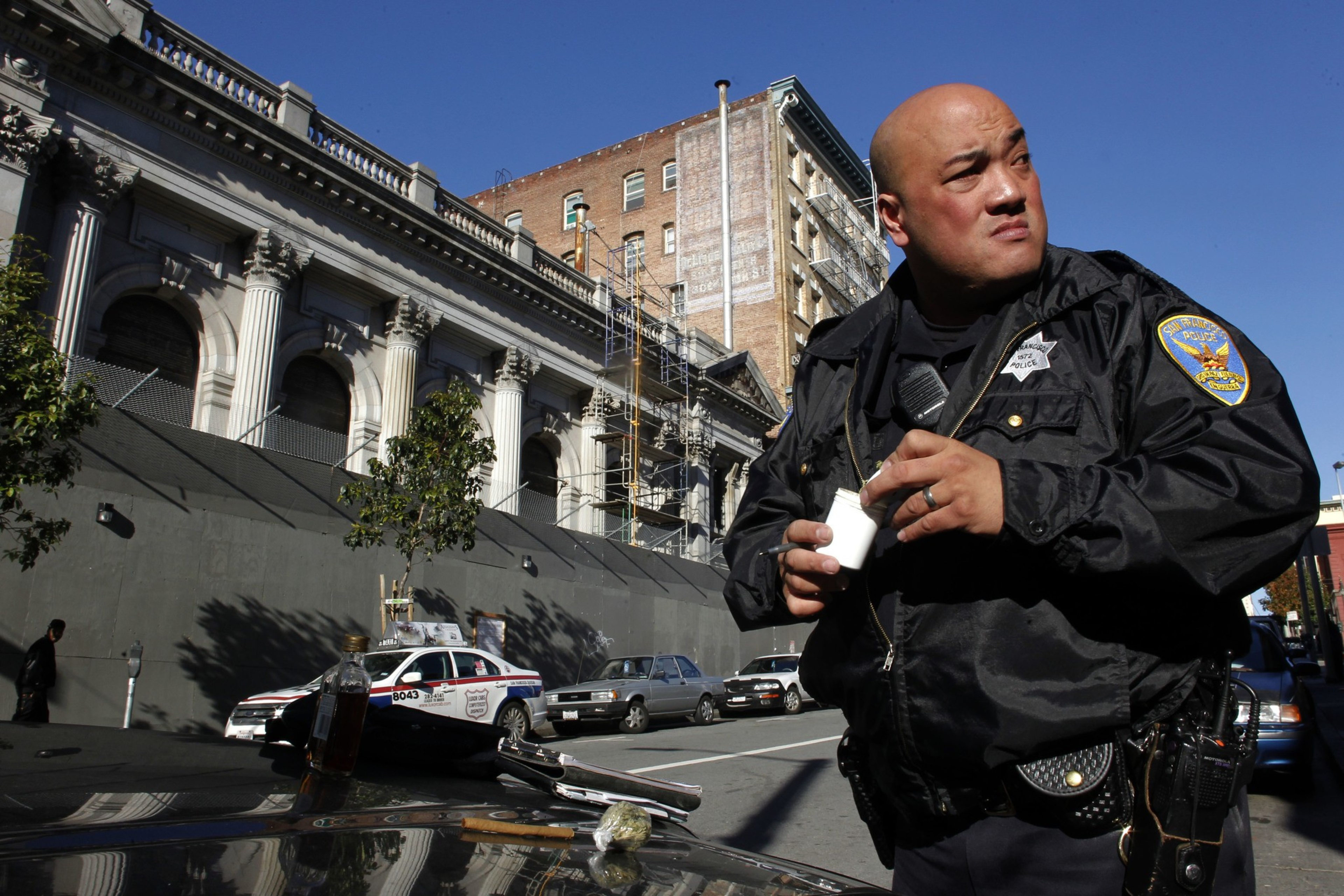 A police officer in uniform stands on a street holding an object, with a building and parked cars behind him under a clear blue sky.