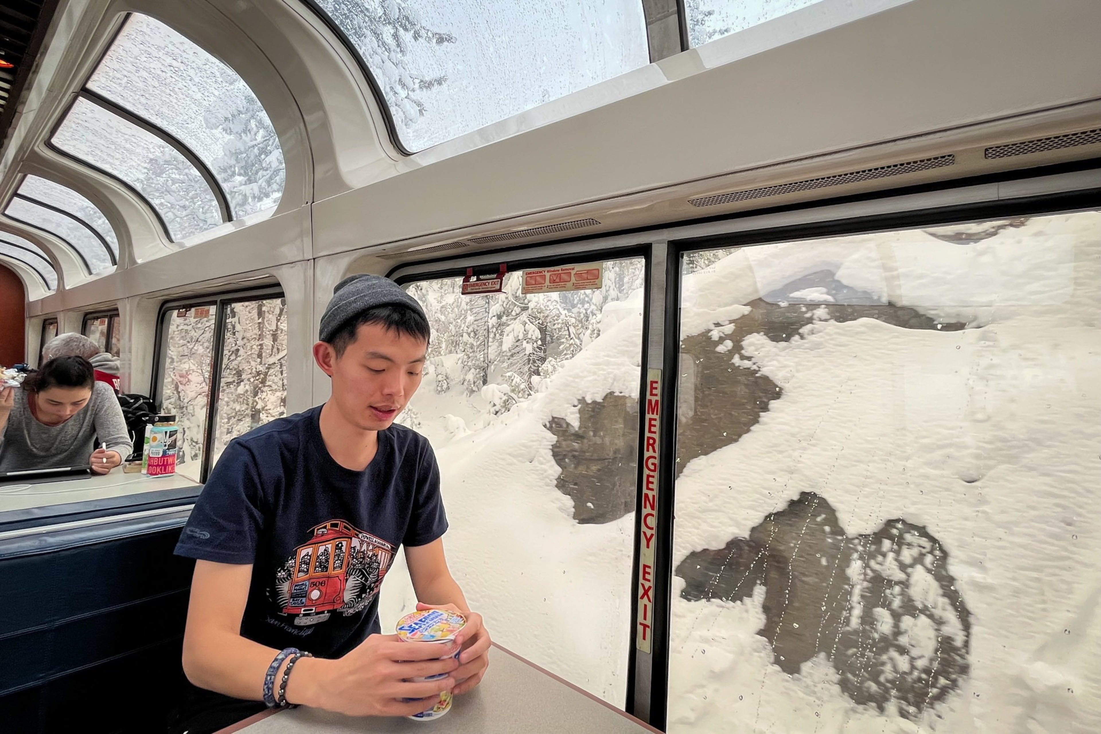 A young man in a beanie and graphic t-shirt sits inside a train car holding a cup, with snow-covered rocks visible through the windows.