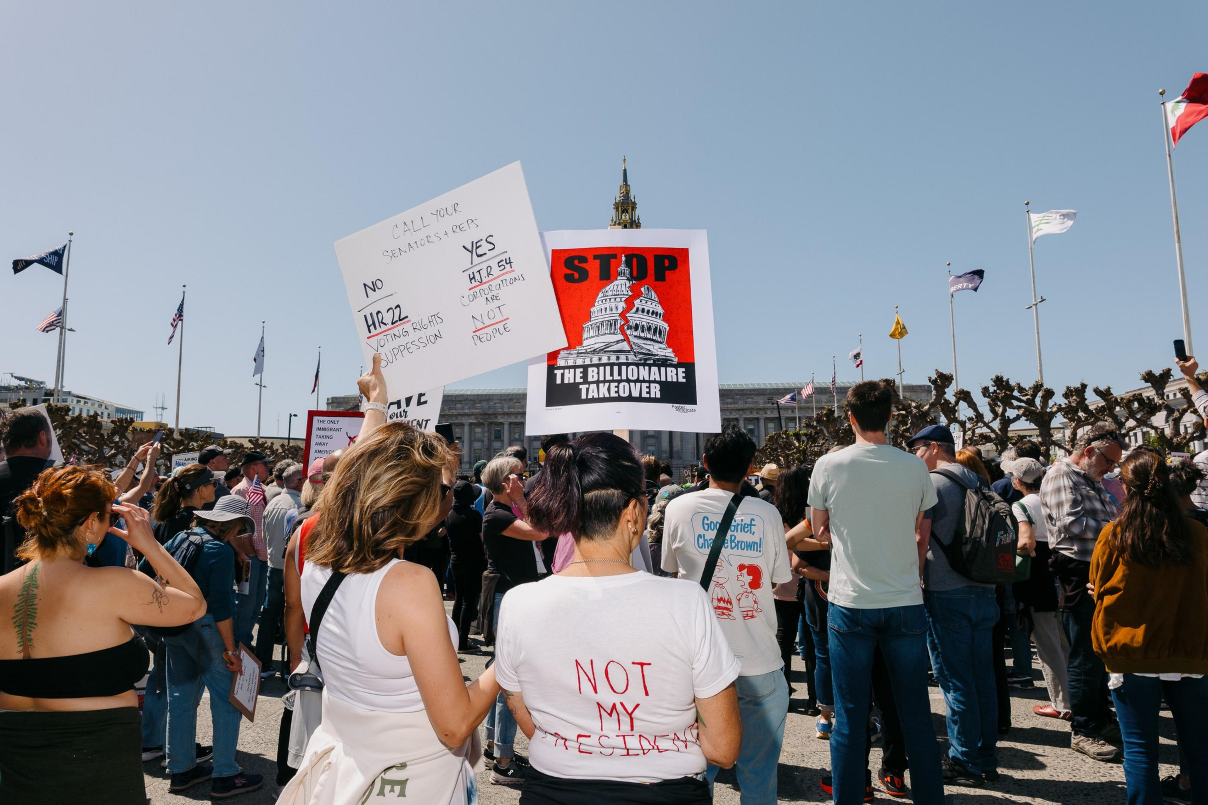 A crowd gathers at a protest with signs against a &quot;billionaire takeover.&quot; Flags wave in the background, and one shirt reads &quot;NOT MY PRESIDENT.&quot;