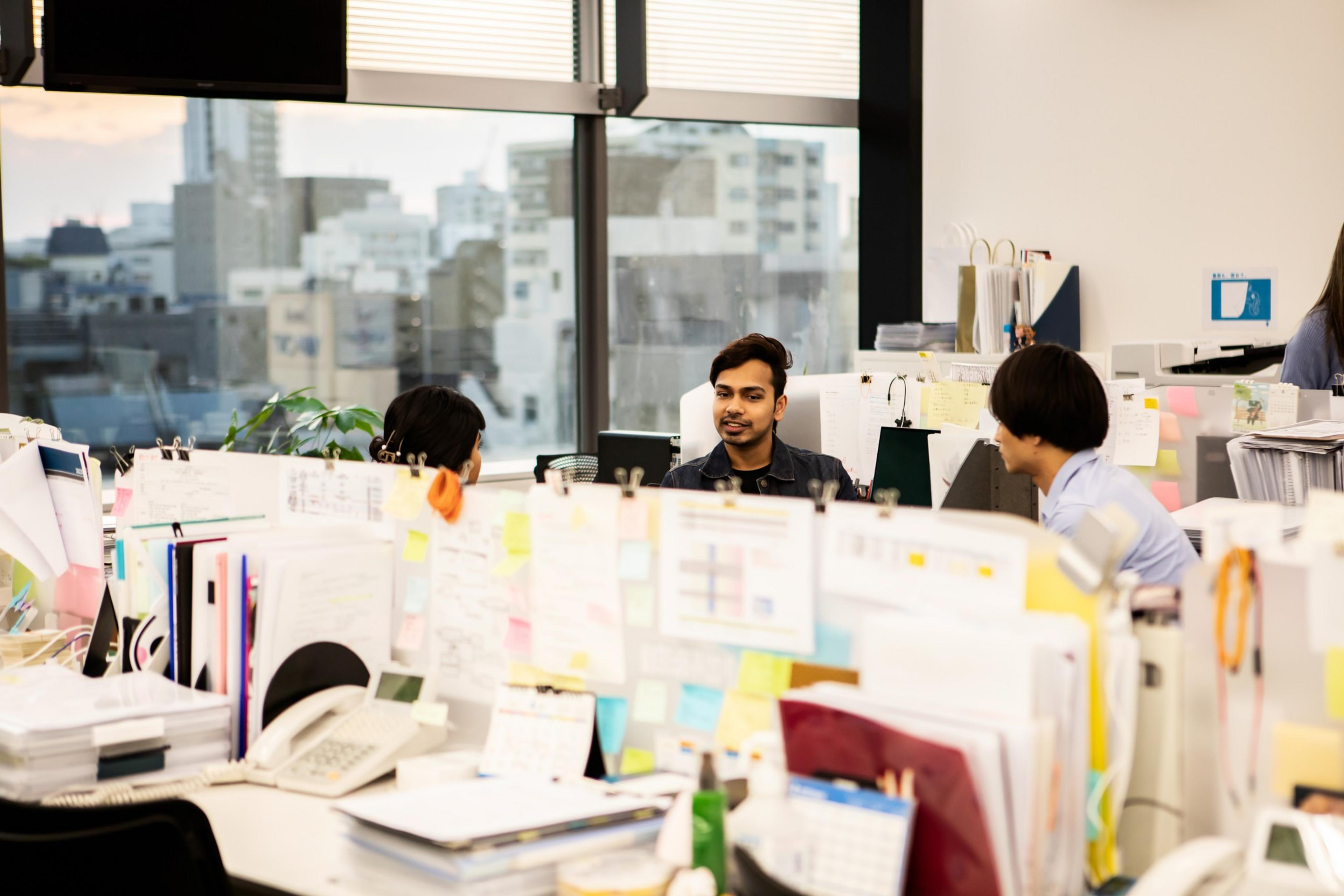 Three people are talking in a bright, cluttered office. The room is filled with papers, folders, and computers. Large windows show a cityscape outside.