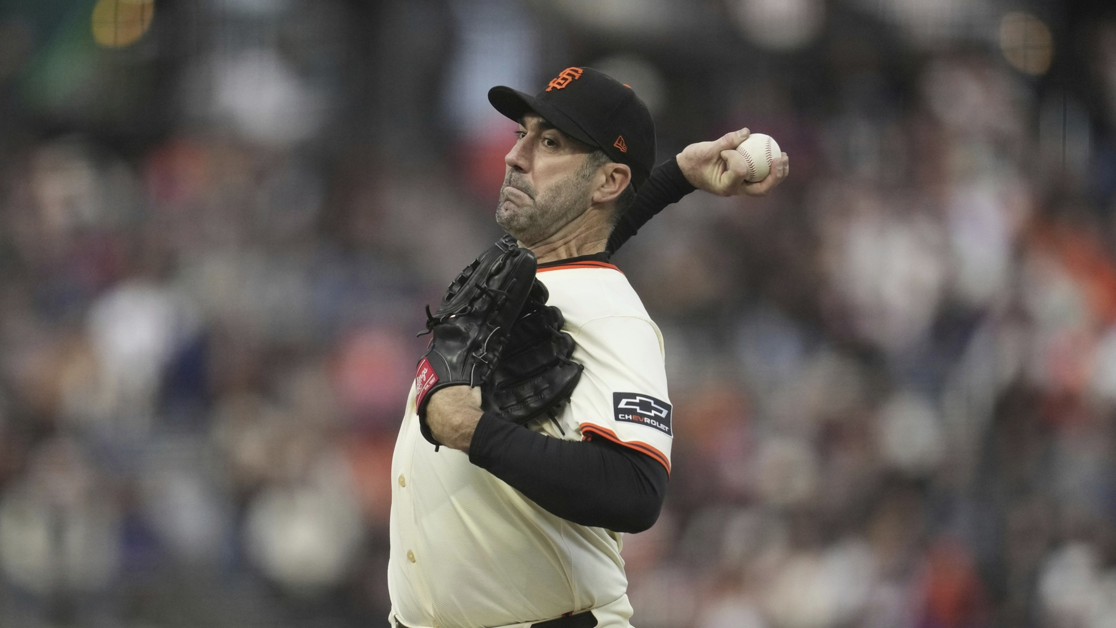 A baseball player in a white uniform with black trim and an orange "SF" cap is mid-pitch, holding a baseball in one hand and wearing a black glove on the other.