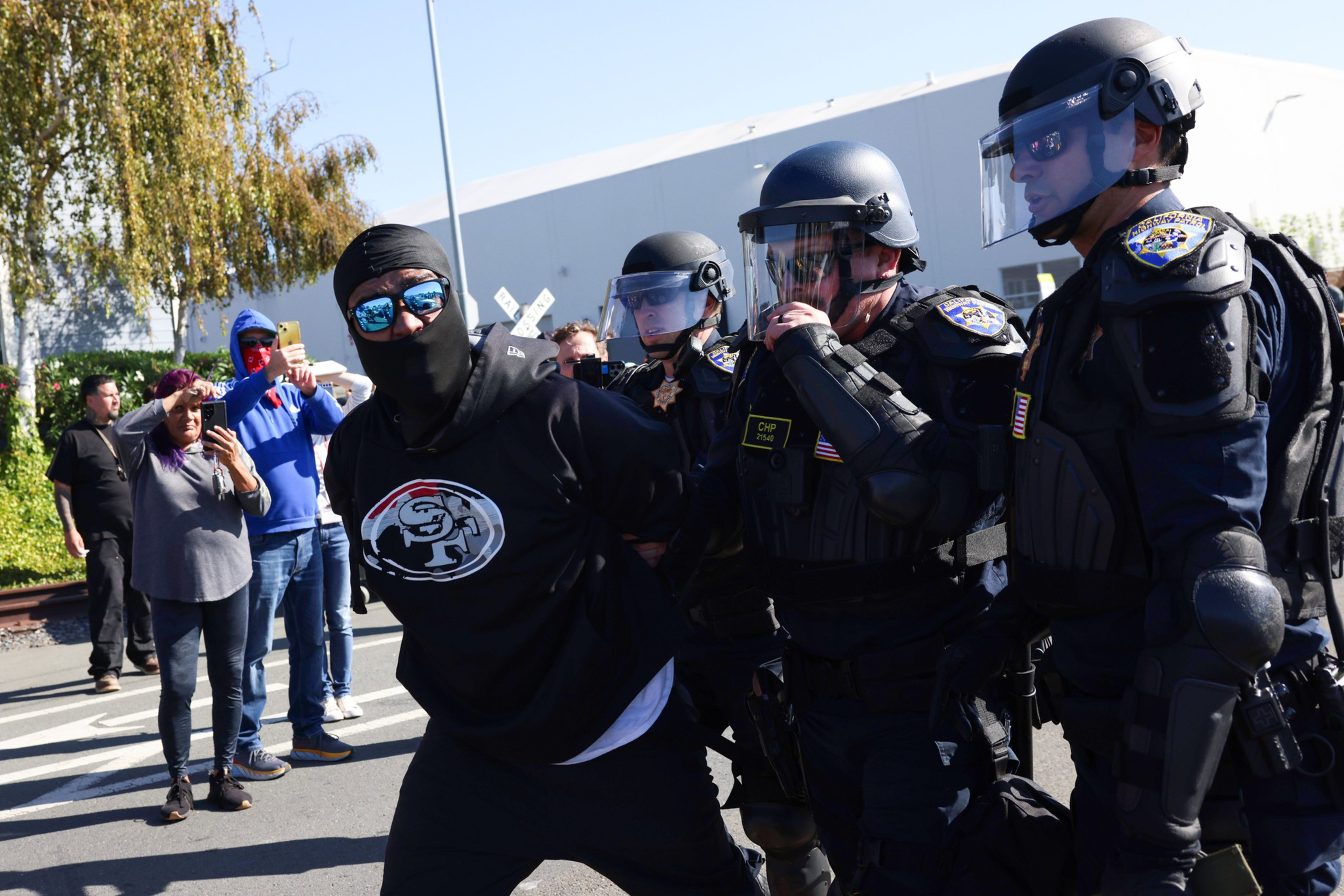 A masked man wearing a black San Francisco 49ers shirt is being detained by police officers in riot gear while bystanders record with their phones.