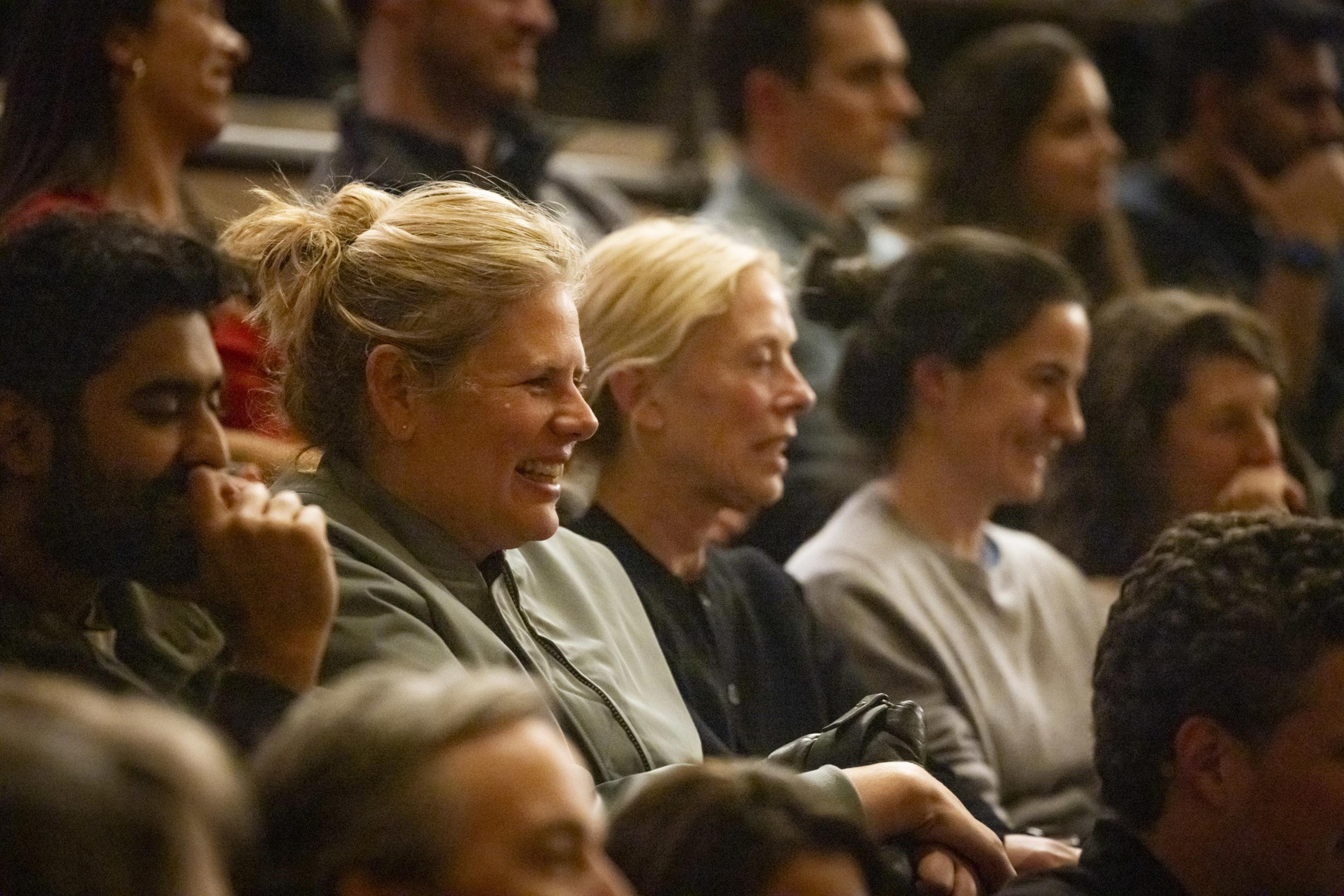 People in an audience are smiling and laughing, seated in rows. The atmosphere appears lively and engaging.