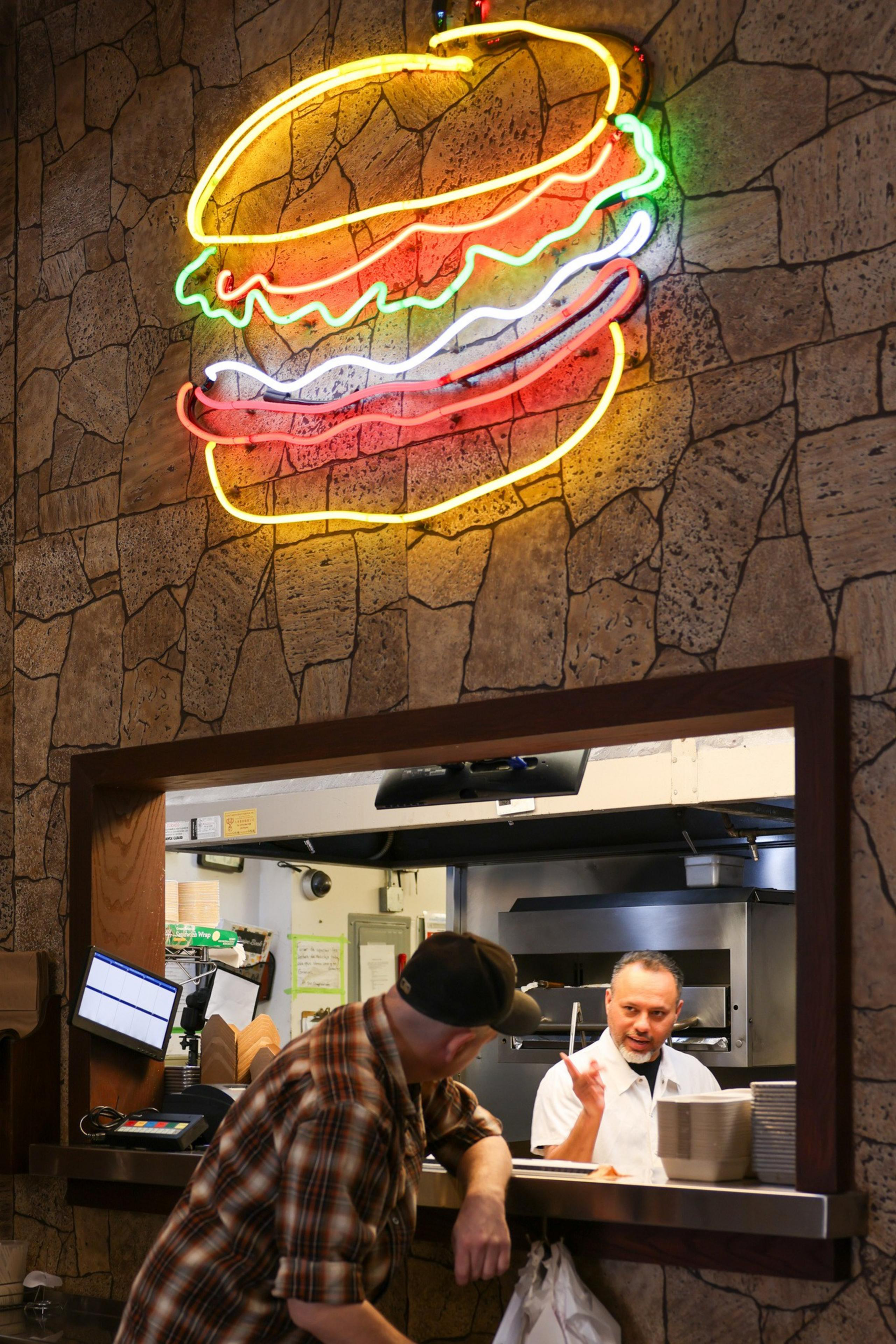 A neon burger sign glows on a stone-patterned wall above a restaurant counter, where a chef speaks with a customer leaning forward in a plaid shirt and cap.