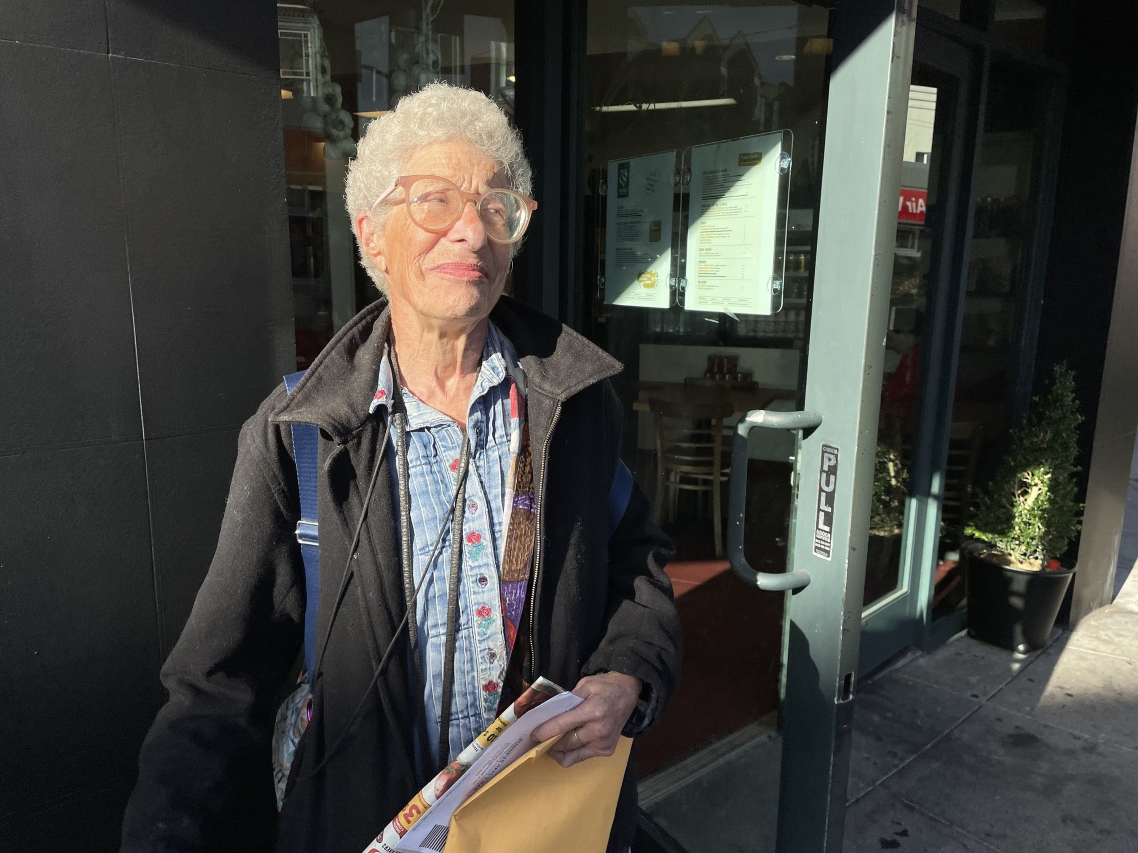 An elderly woman stands holding mail in front of a neighborhood restaurant on a street corner in San Francisco.