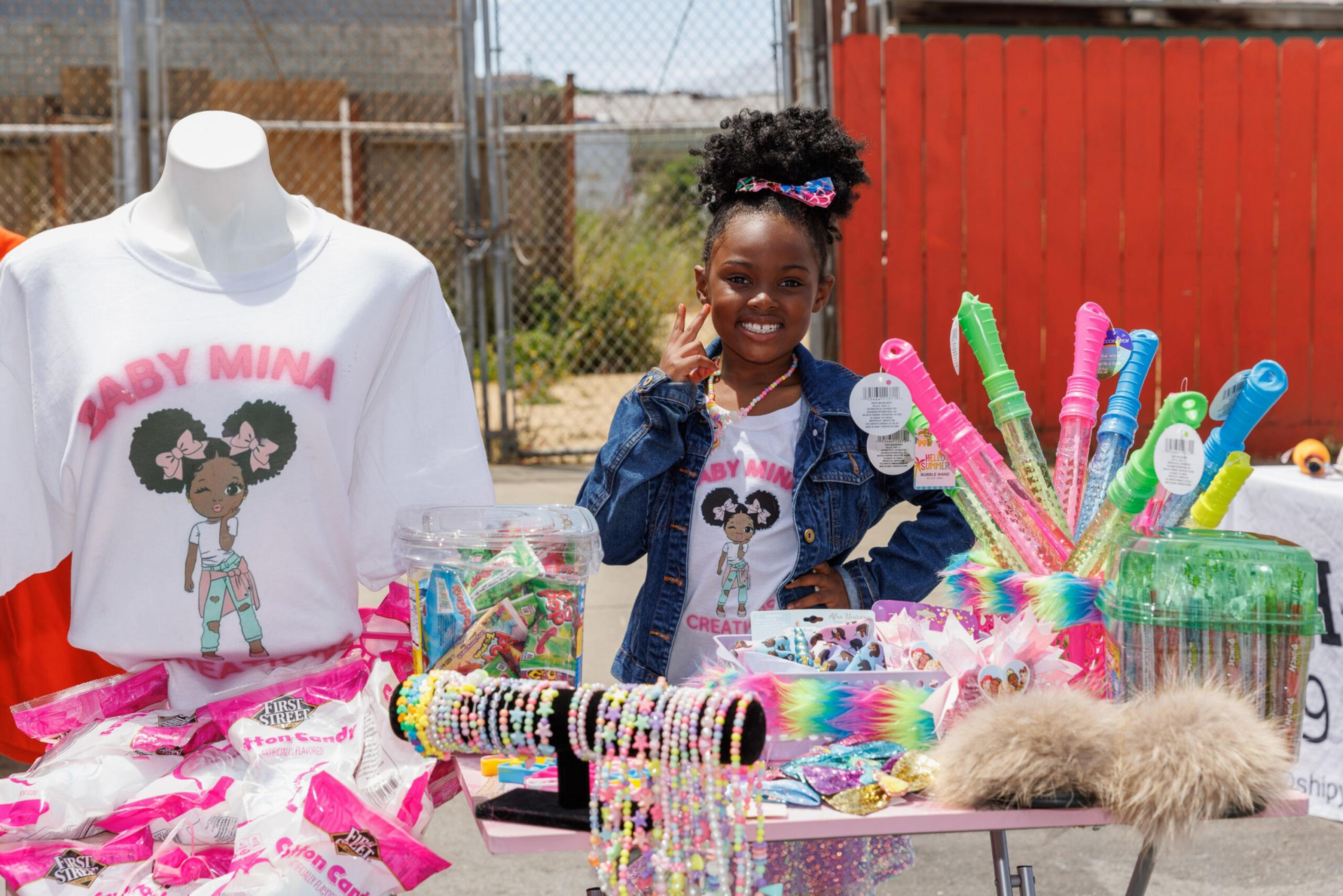 A smiling girl stands by a table with colorful merchandise and a shirt with &quot;BABY MINA&quot; printed on it.