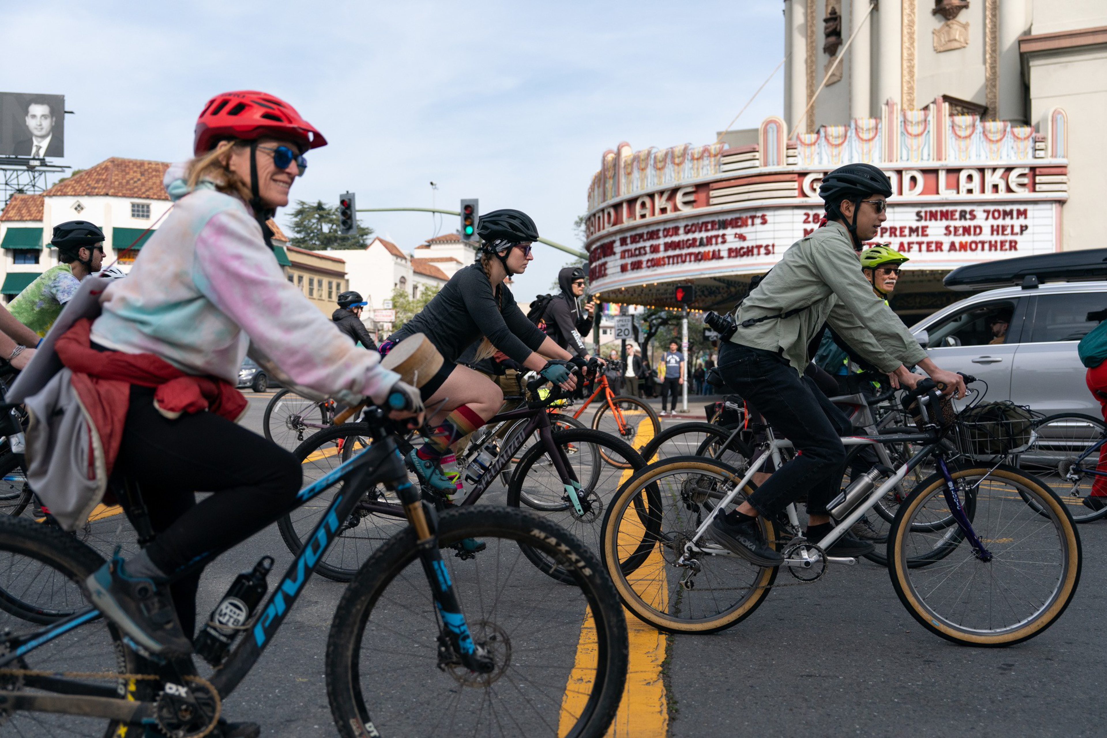 Several cyclists wearing helmets ride through a city street intersection near a theater marquee that reads "GRAND LAKE."