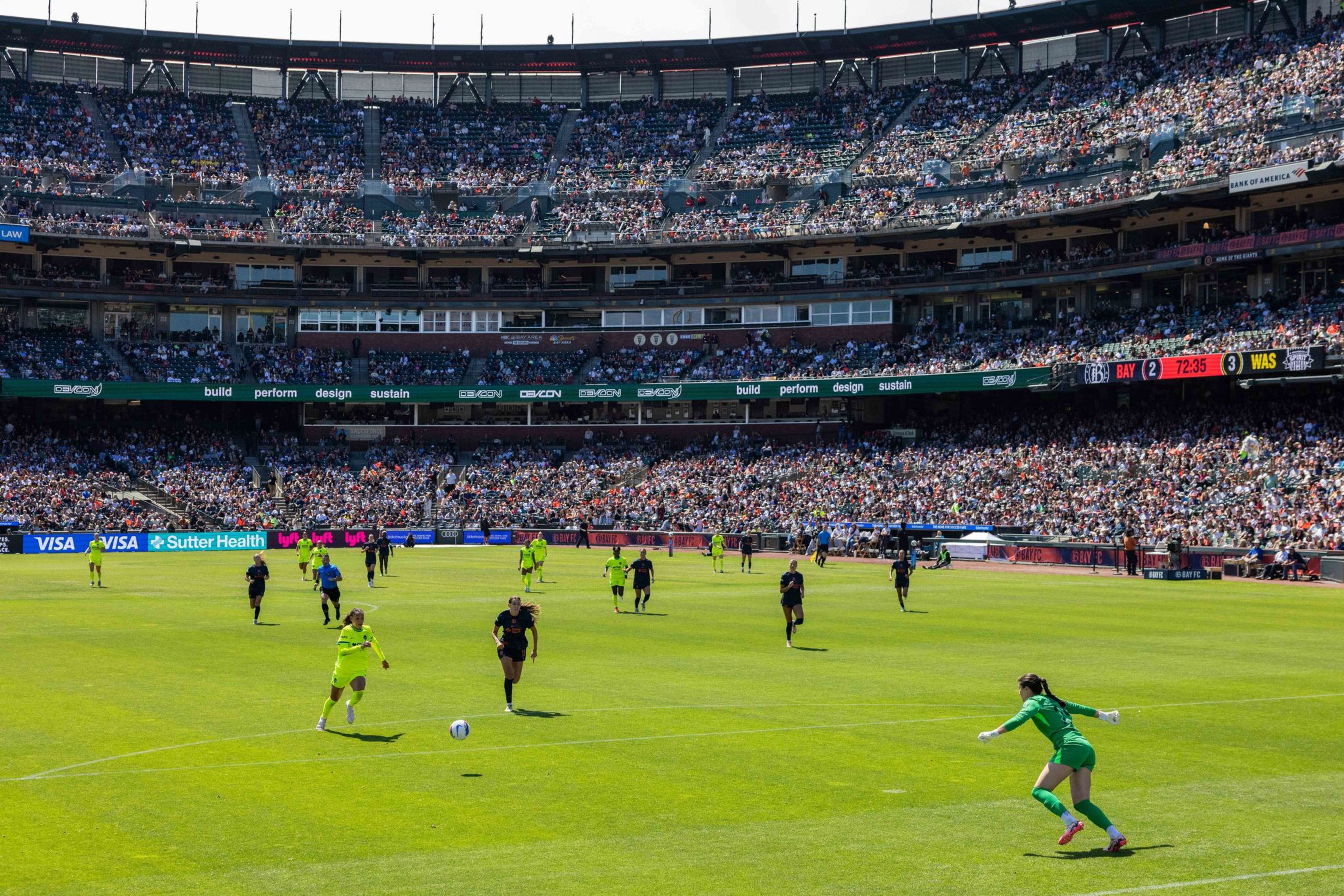 A soccer game is in progress with a player in yellow running toward the ball and a goalkeeper in green ready to defend, while a large crowd fills the stadium.