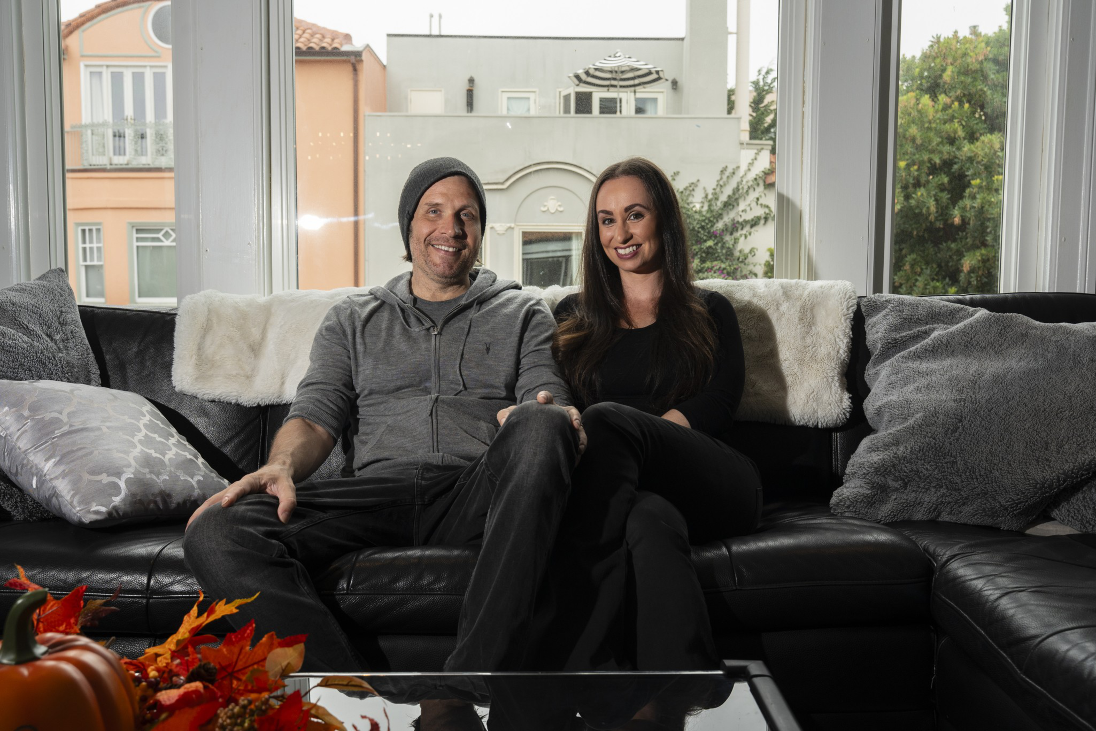 A man in a gray hoodie and beanie and a woman with long brown hair, both smiling, sit closely on a black leather couch with cozy pillows.