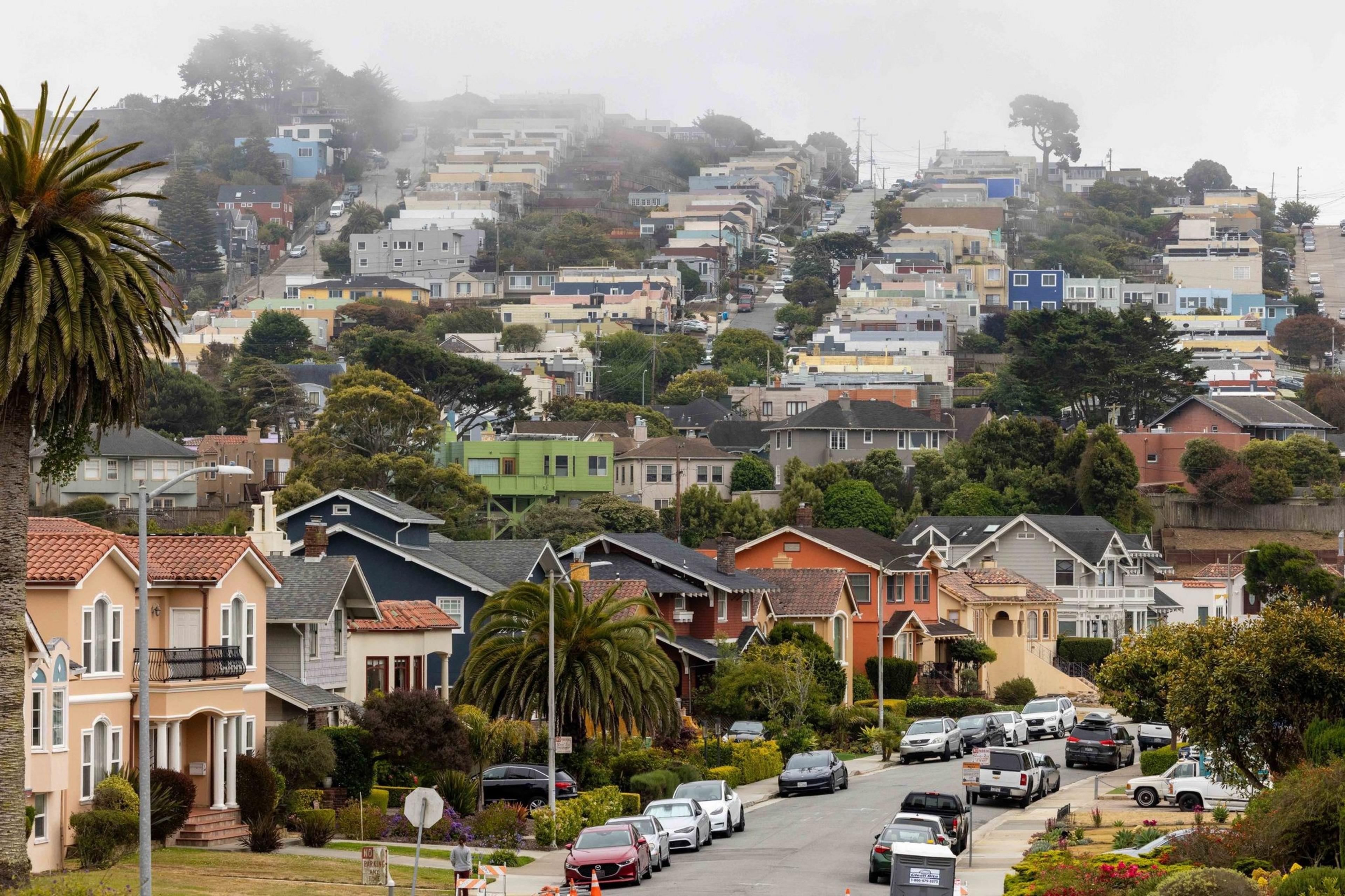 The image shows a hilly neighborhood with colorful houses and lush greenery. Cars line the street, and a foggy sky hovers above the distant hills.