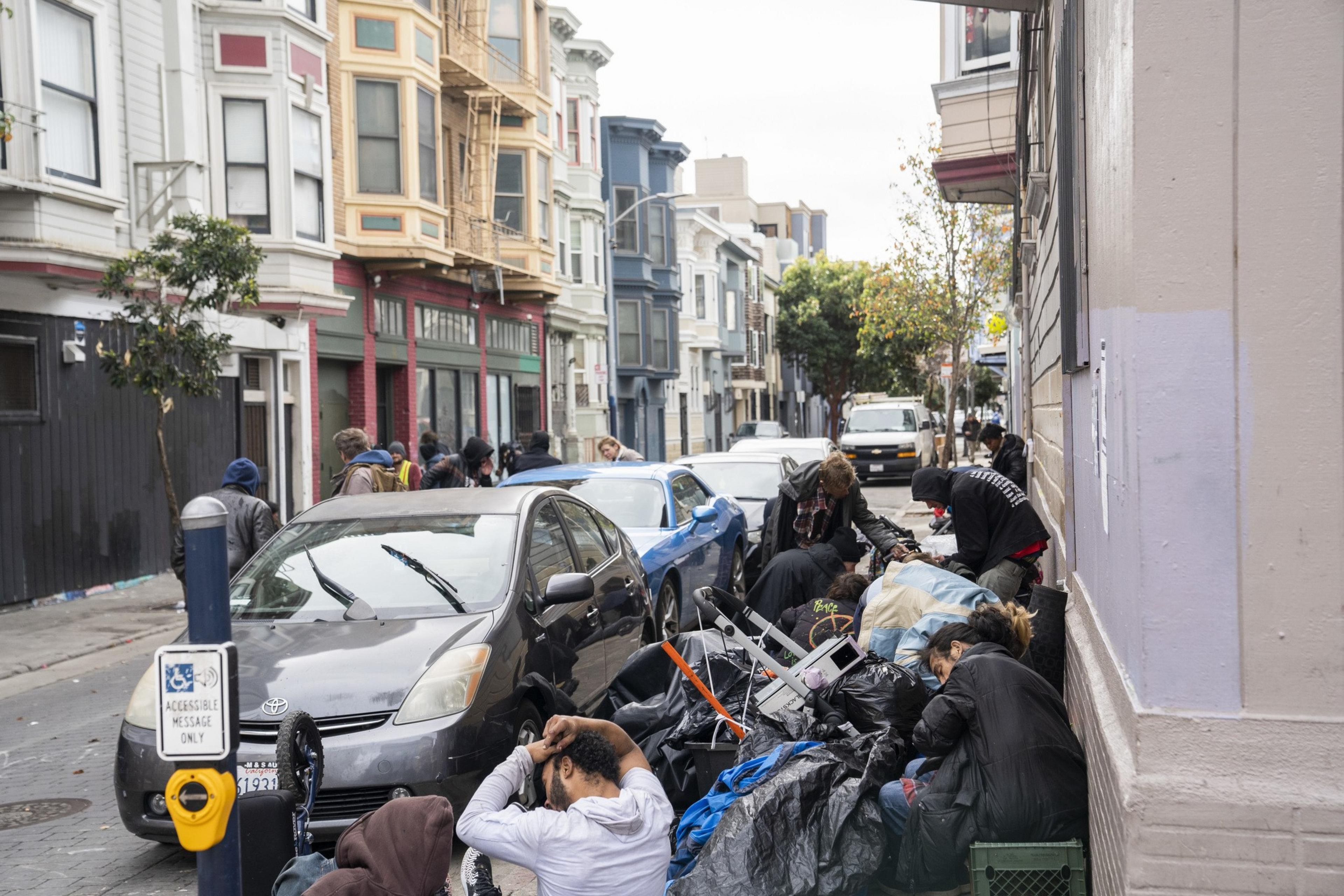 People are gathered on a street cluttered with bags and belongings, beside parked cars and colorful buildings, depicting an urban scene.