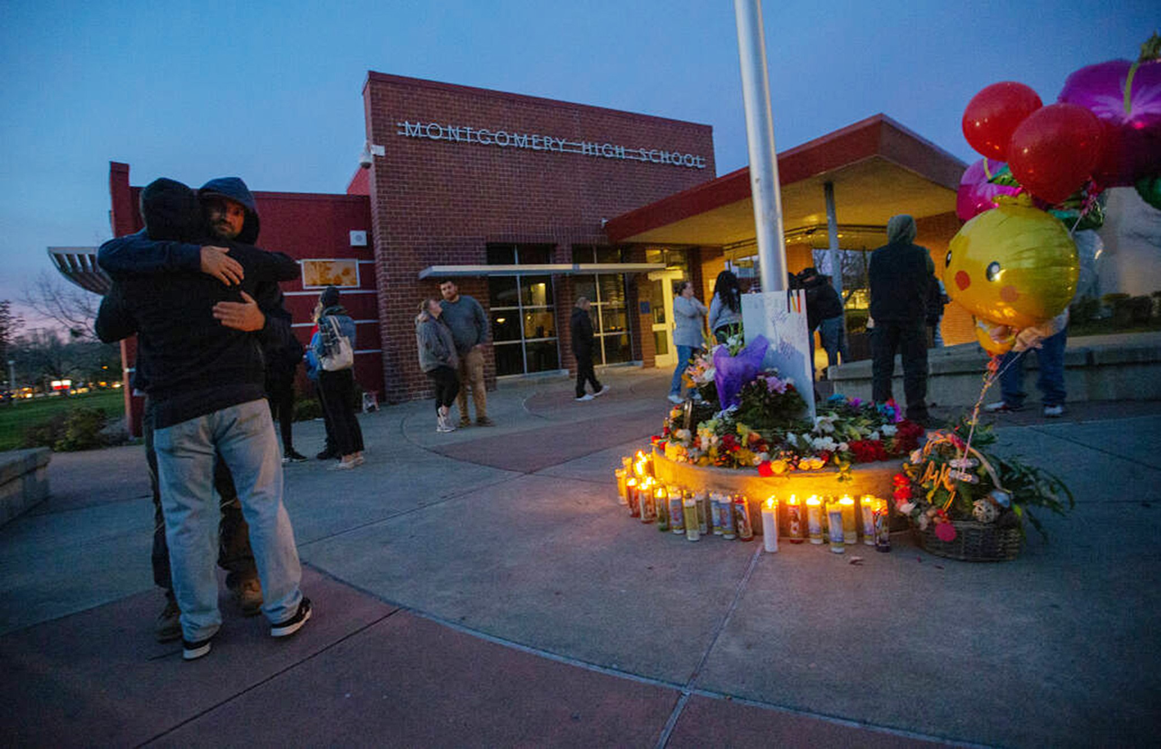 People gather outside Montgomery High School at dusk around a memorial with lit candles, flowers, and balloons, while two individuals embrace.