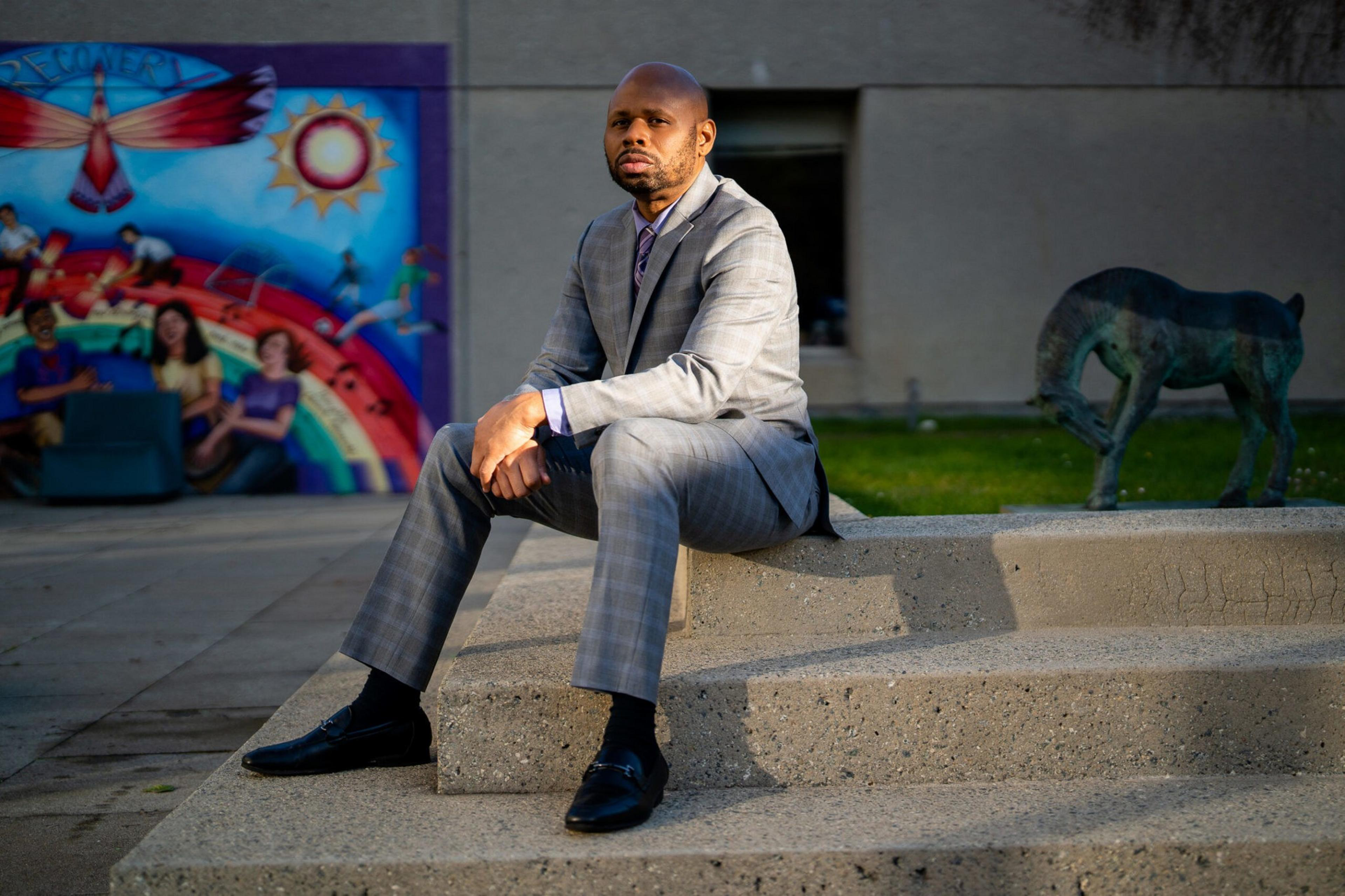 A man in a gray plaid suit sits on concrete steps with a colorful mural and an abstract animal sculpture behind him.