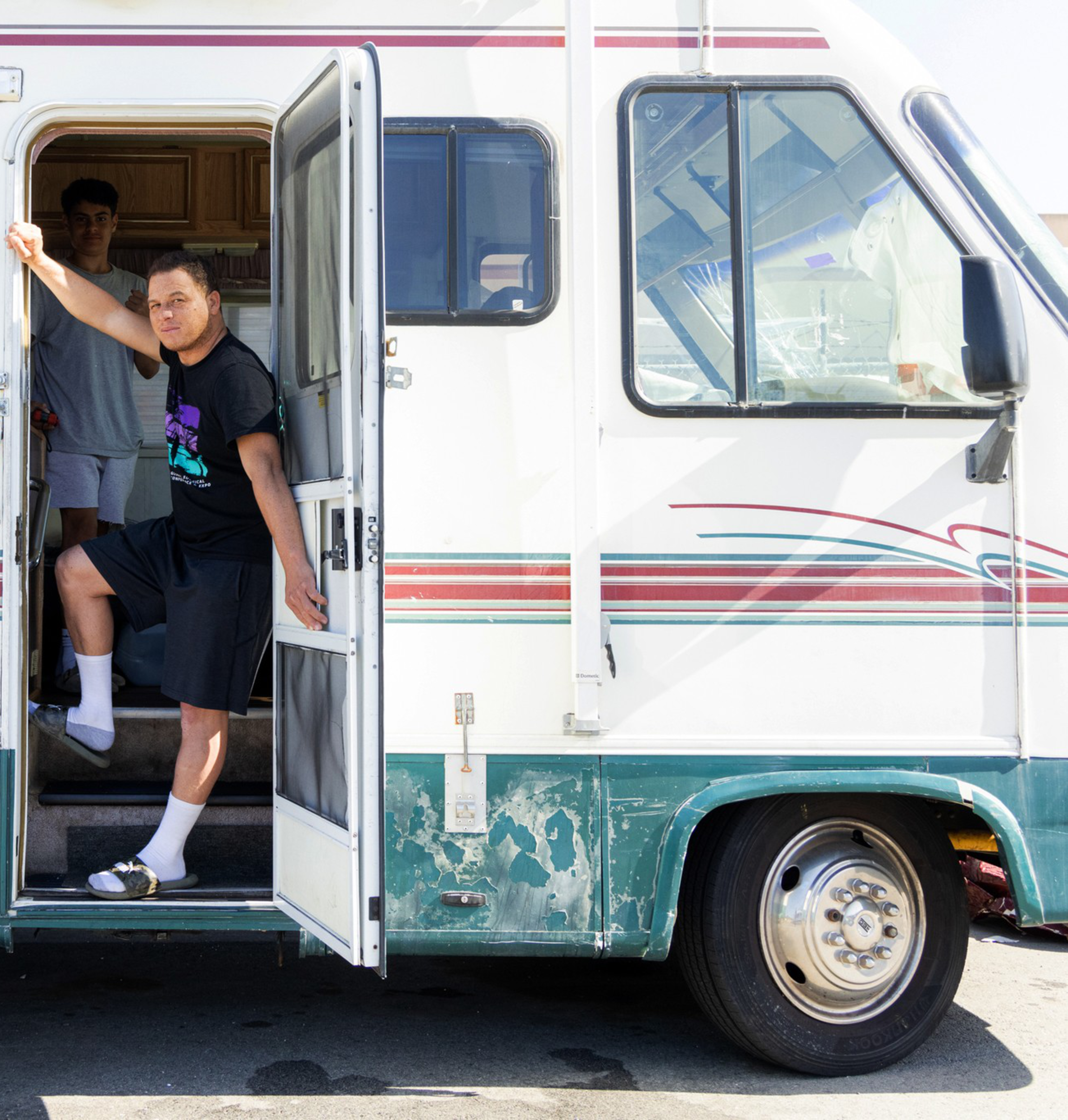 Two people are at the open door of an old RV; one stands outside, leaning on the door frame, while the other is inside, partially visible.