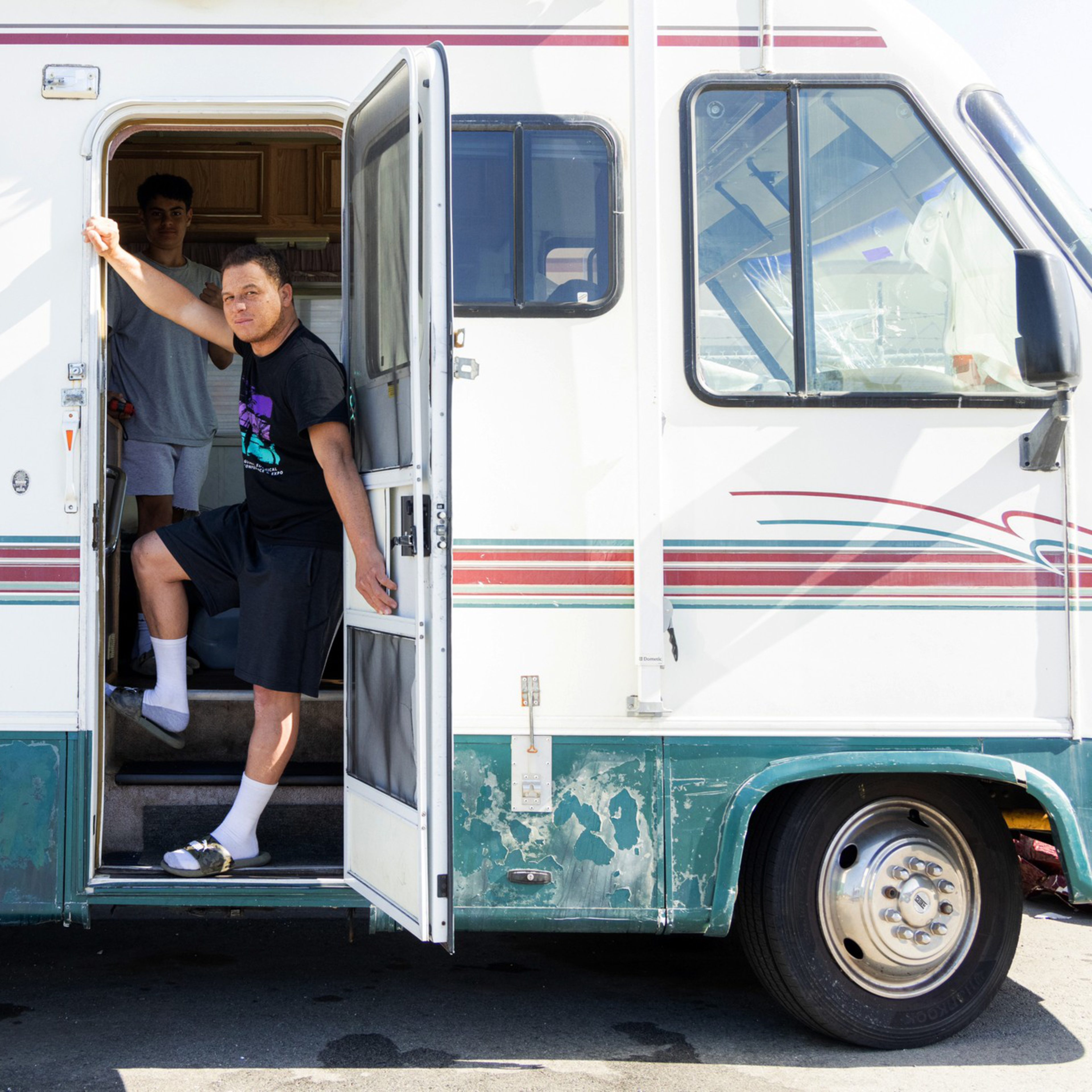 Two people are at the open door of an old RV; one stands outside, leaning on the door frame, while the other is inside, partially visible.