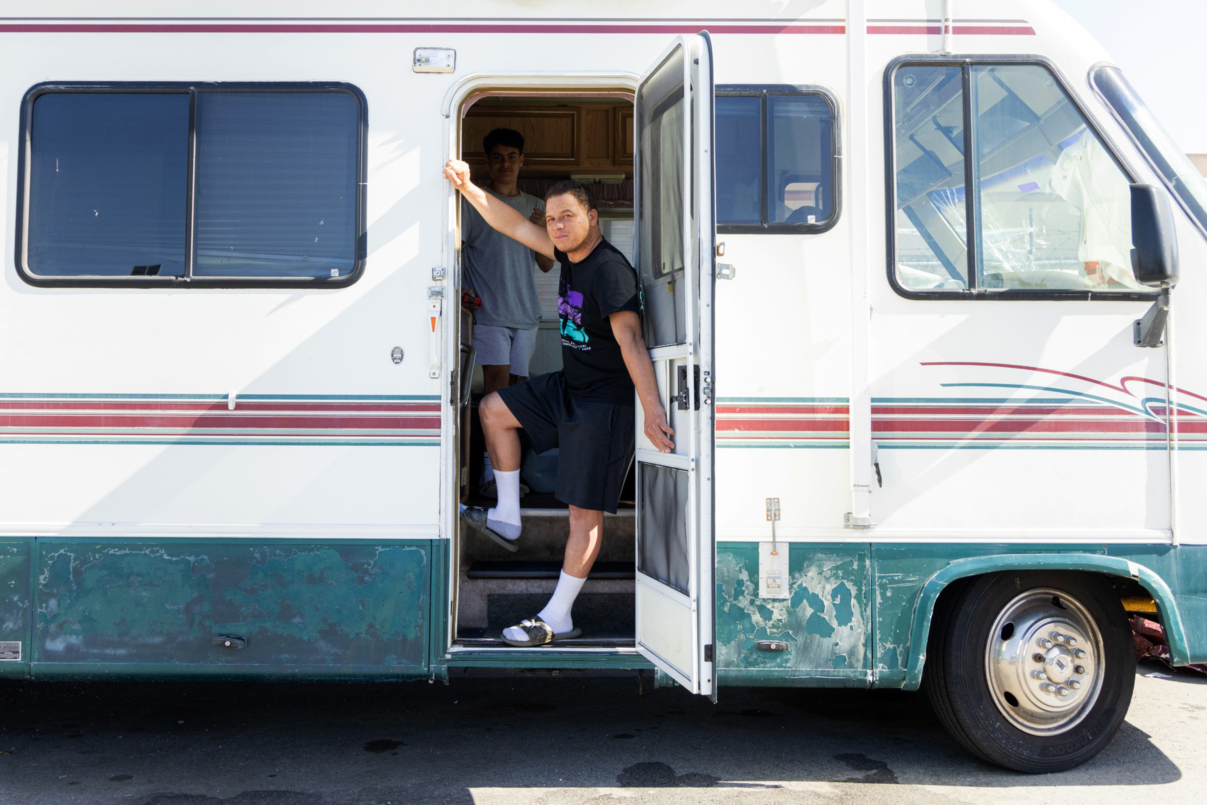Two people are at the open door of an old RV; one stands outside, leaning on the door frame, while the other is inside, partially visible.