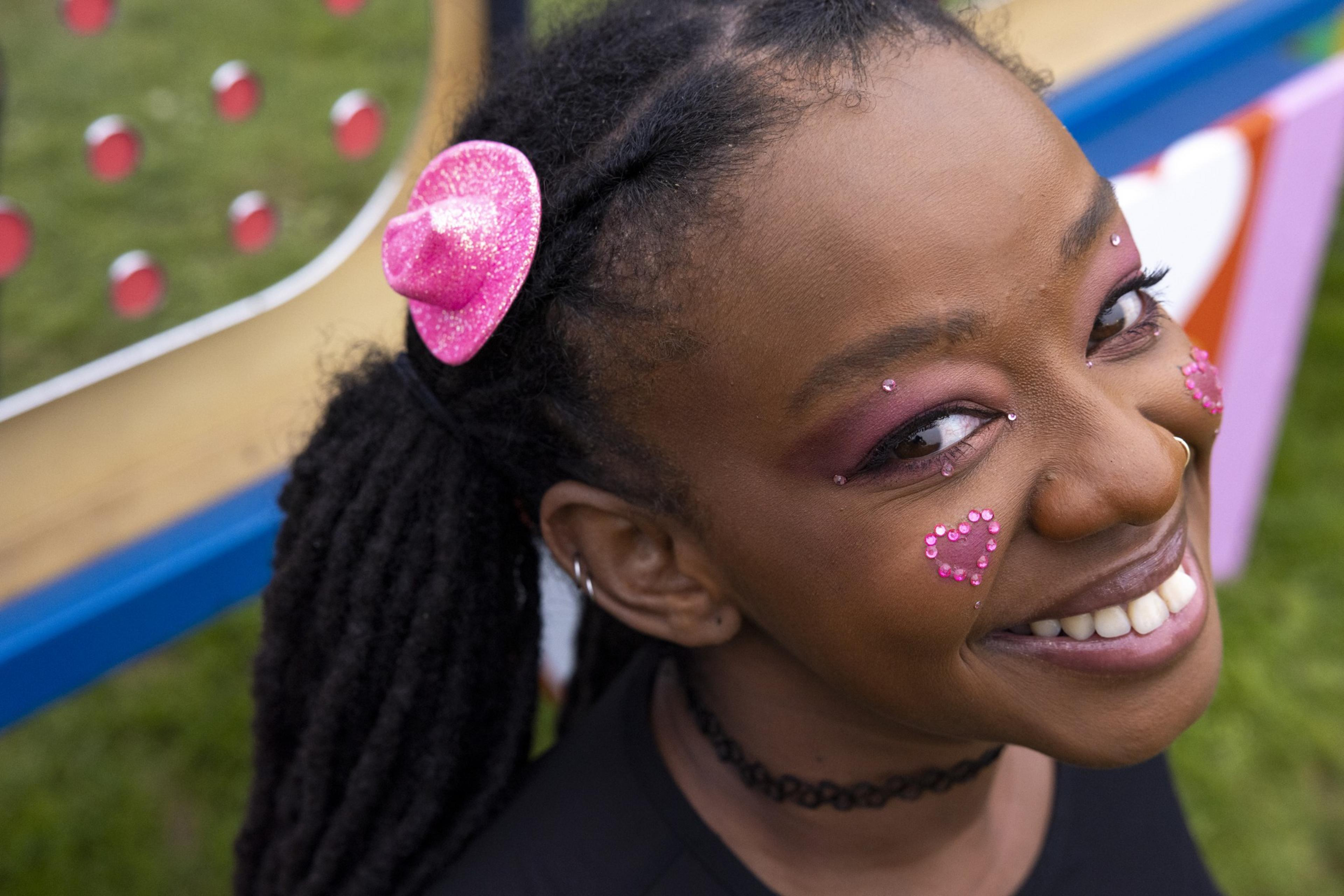 A smiling person with a pink glittery hat accessory, pink eye makeup, and heart-shaped face gems. The background shows an outdoor setting with colorful, blurred objects.