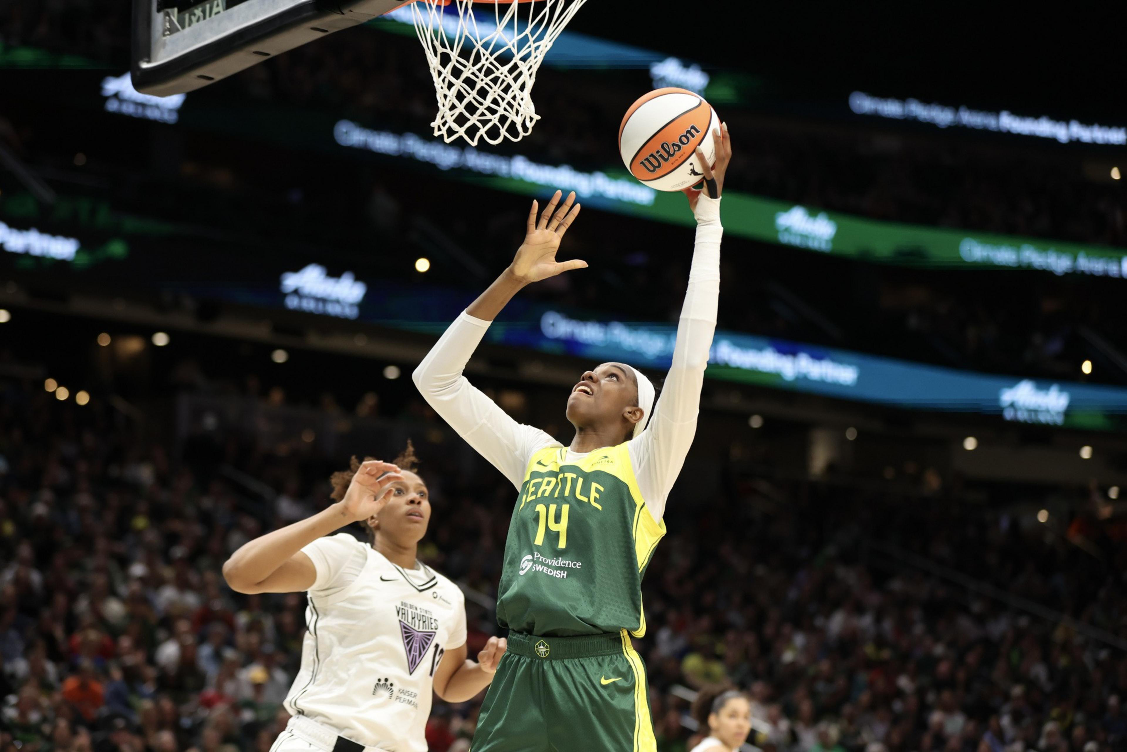 A basketball player in a green Seattle jersey leaps to shoot the ball near the hoop, while a defender in a white jersey closely guards her.