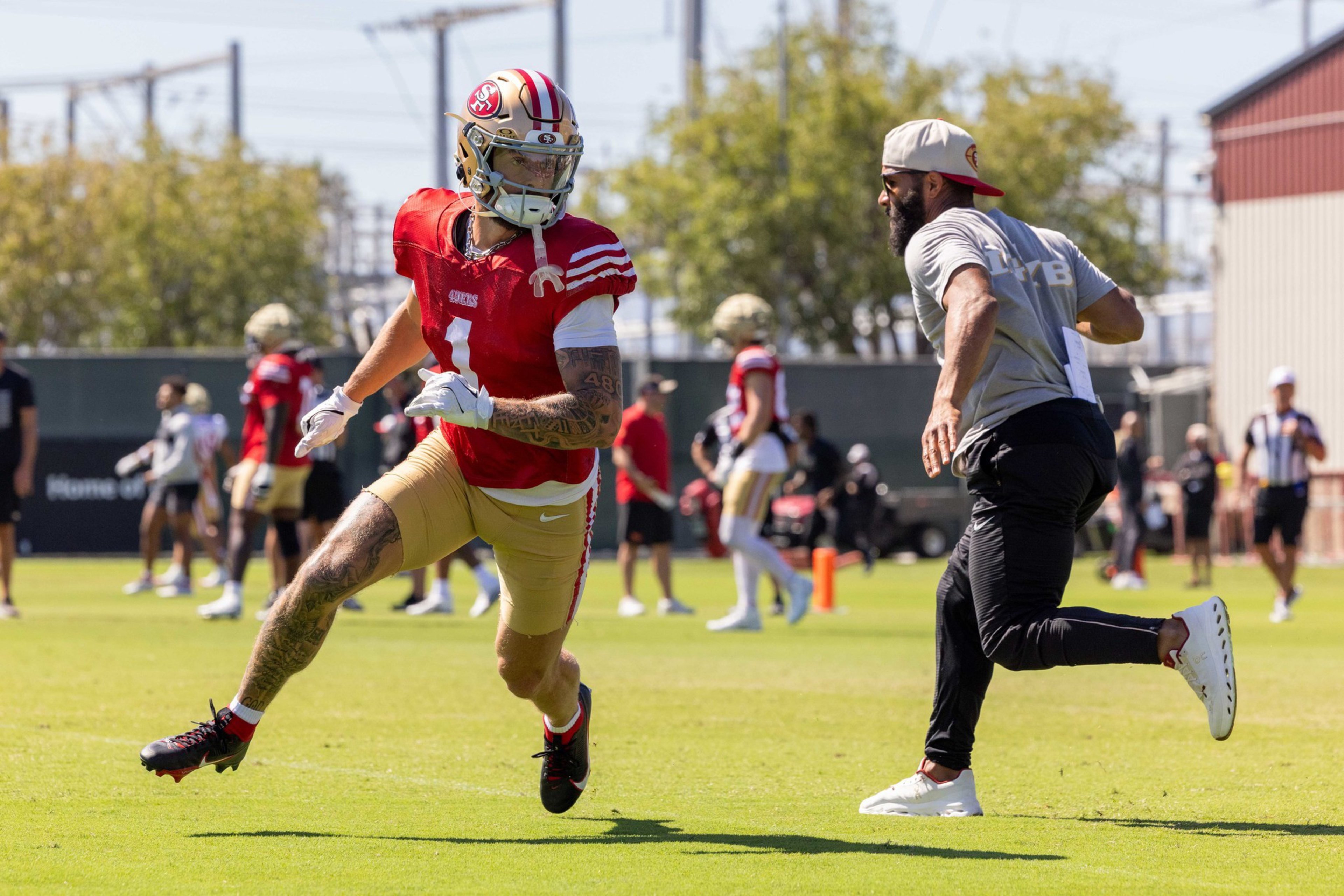A football player in a red jersey and gold pants runs on grass as a man in a gray shirt and black pants chases him during practice.