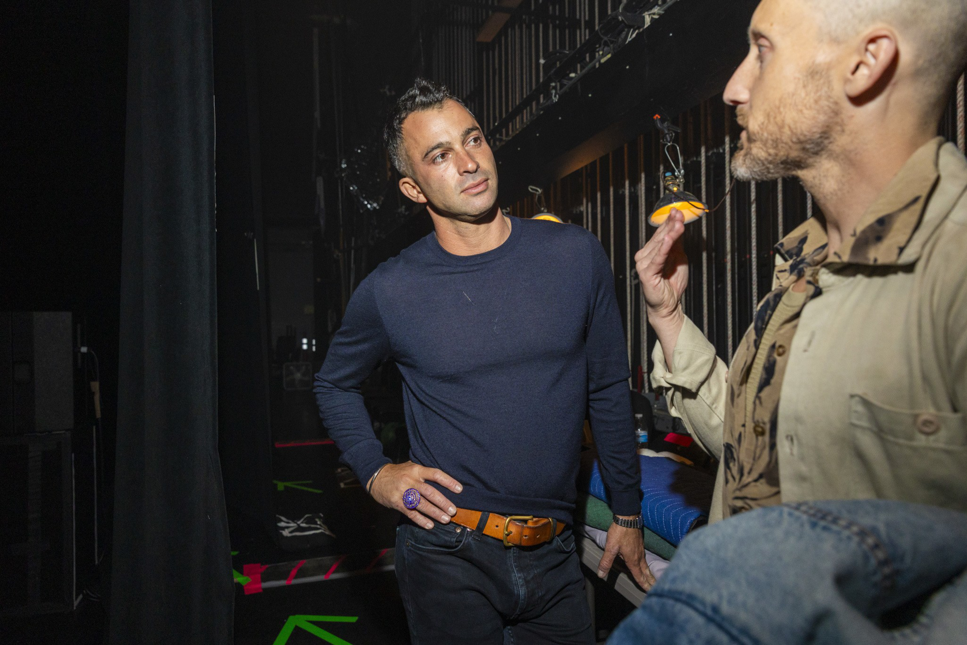 A man in a navy shirt and orange belt listens attentively to another man holding an orange slice in a dimly lit backstage area.