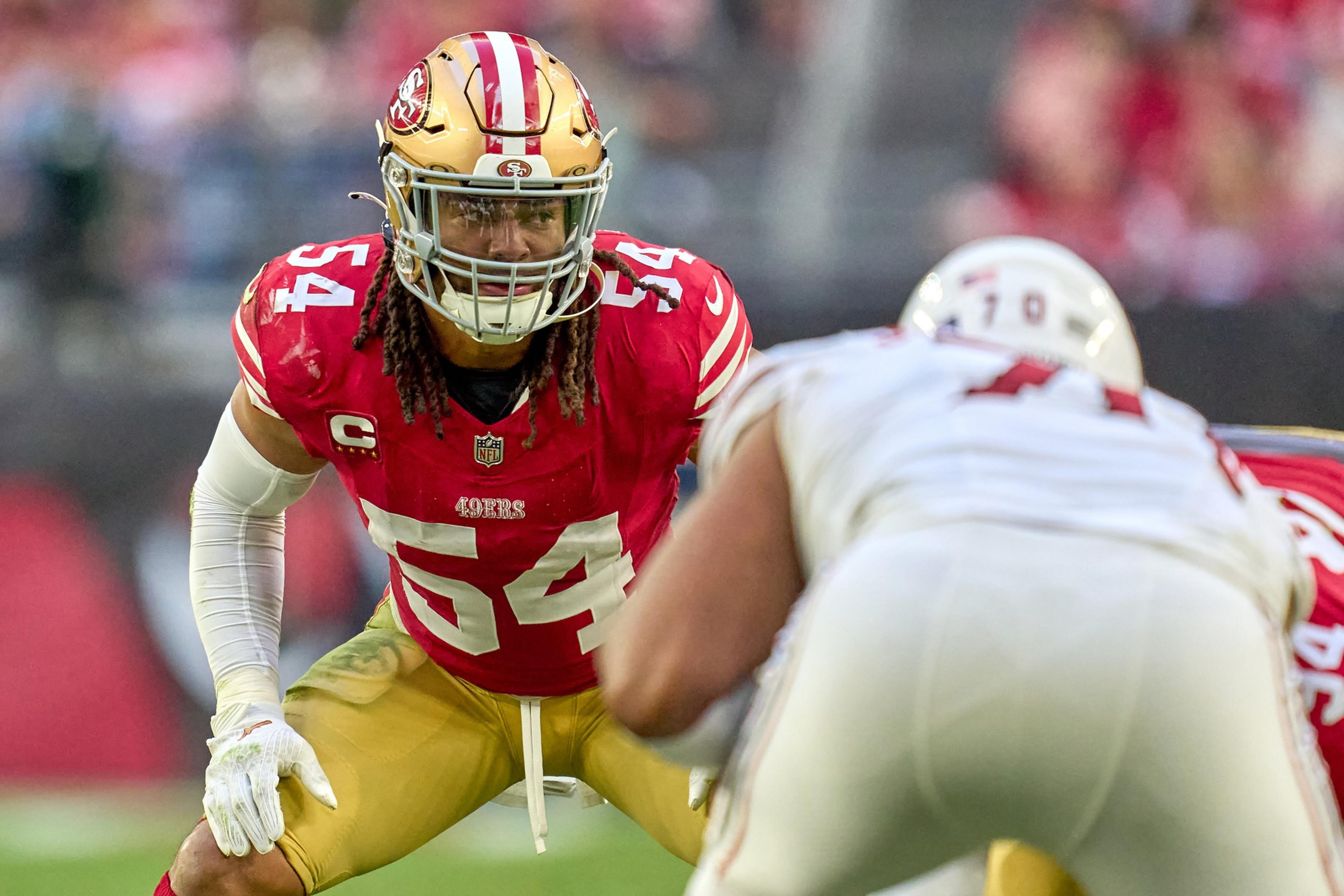 A football player in a red and gold uniform with number 54 crouches in a ready position, facing an opponent in a white uniform during a game.
