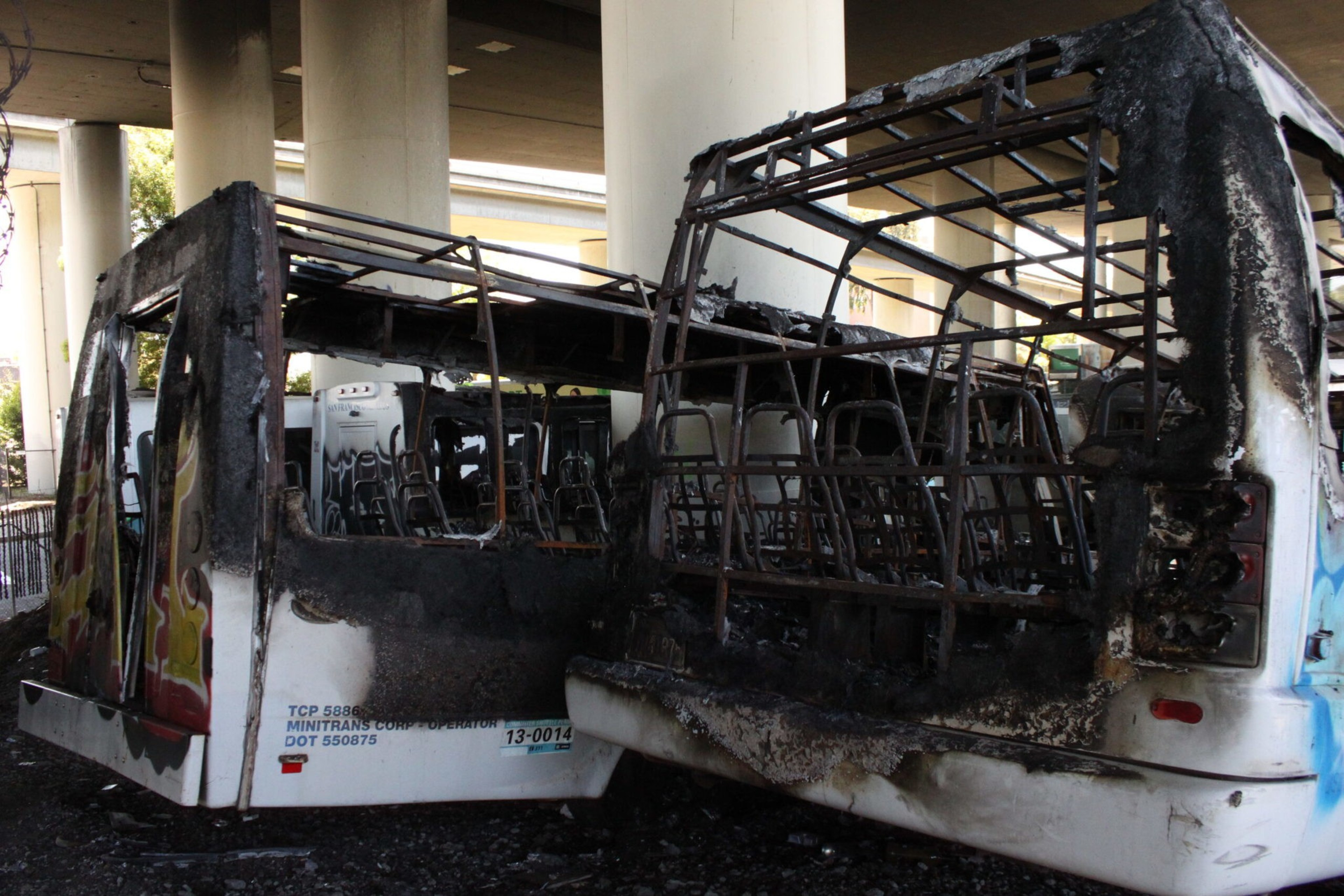 Two severely burned and gutted bus shells with exposed metal frames are parked under a highway overpass.