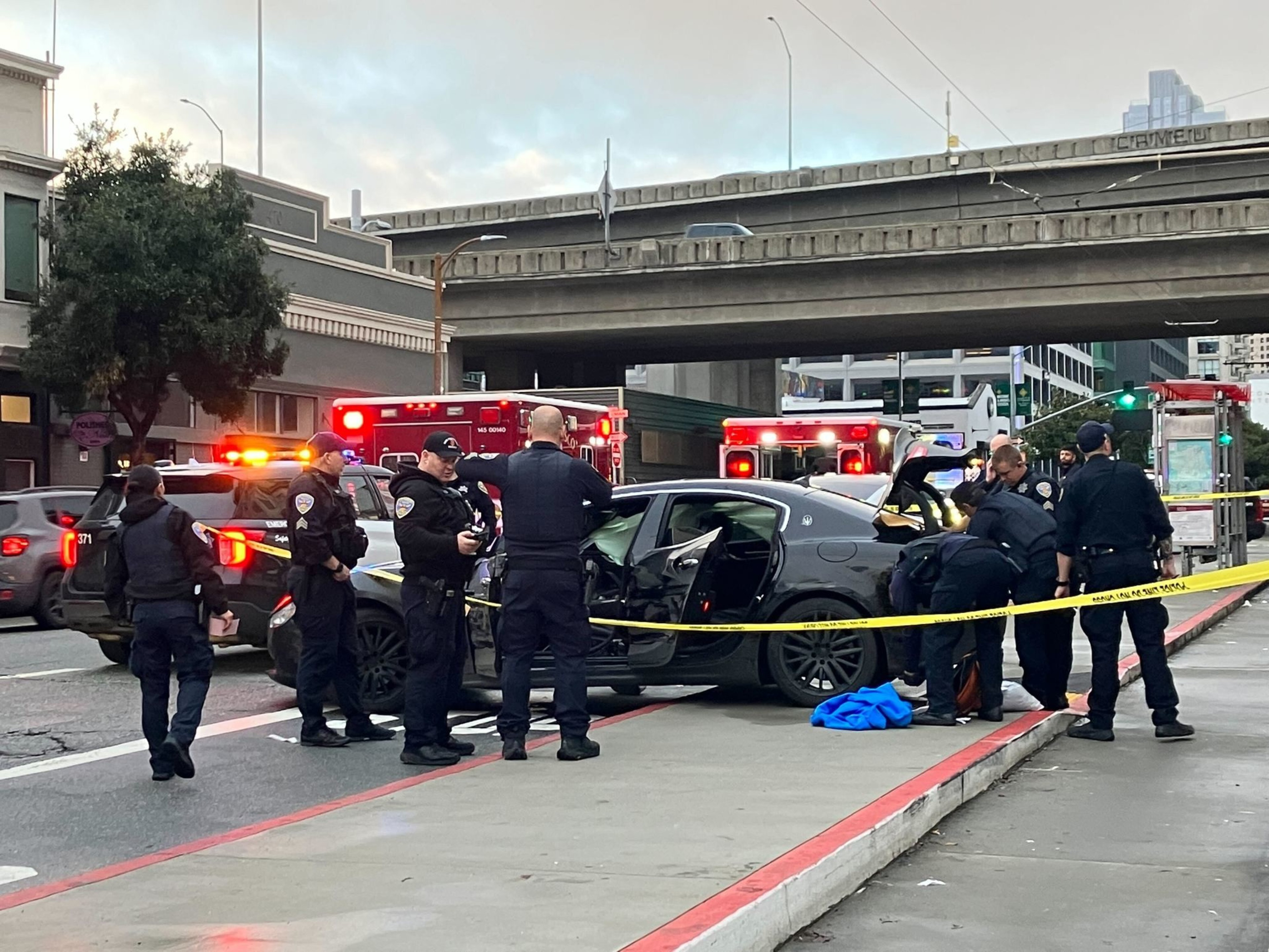 Emergency responders are at a street scene with a black car, police officers, and flashing lights under an overpass.
