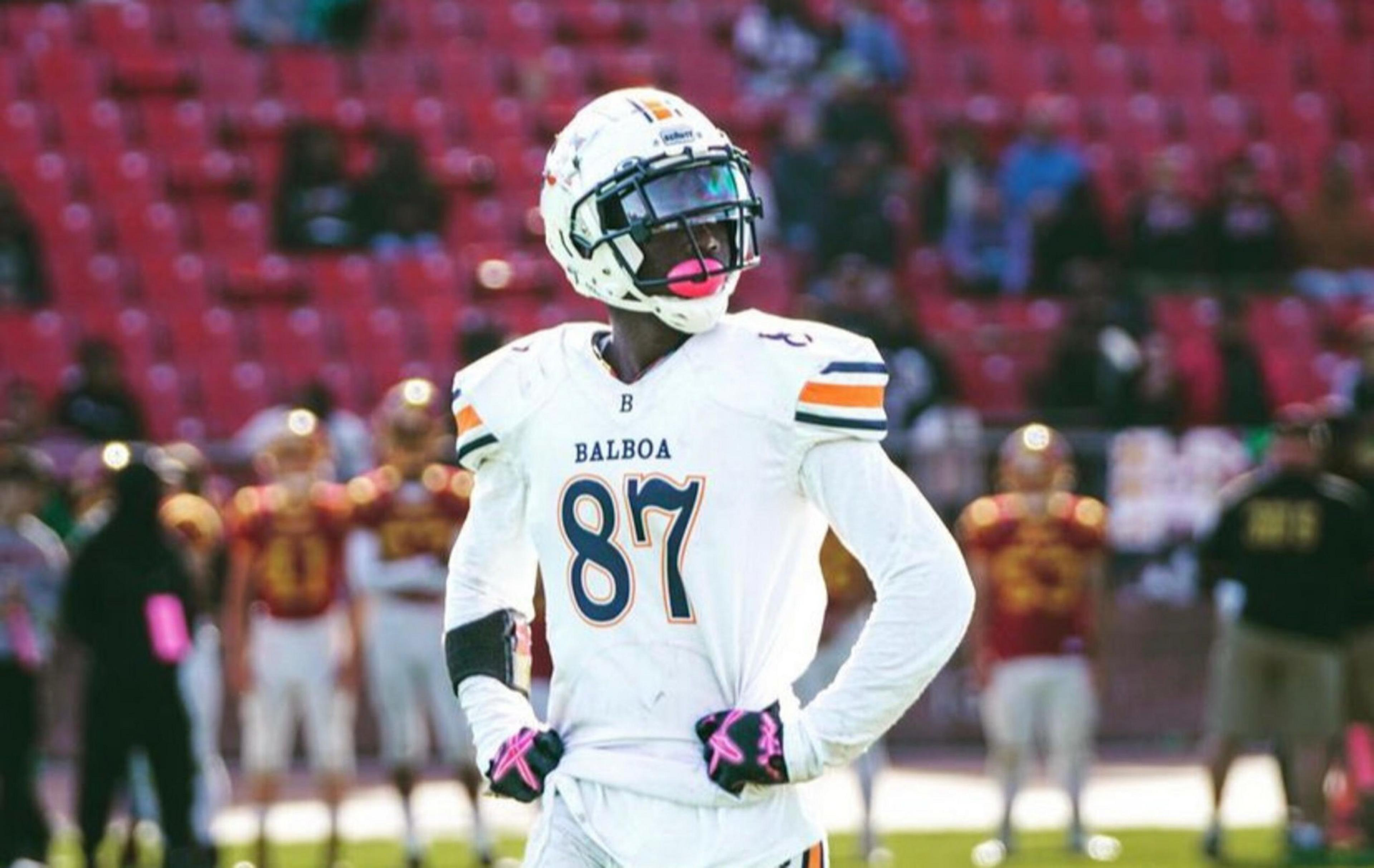 A football player wearing a white Balboa jersey with number 87 stands with hands on hips, looking to the side on a field with blurred spectators behind.
