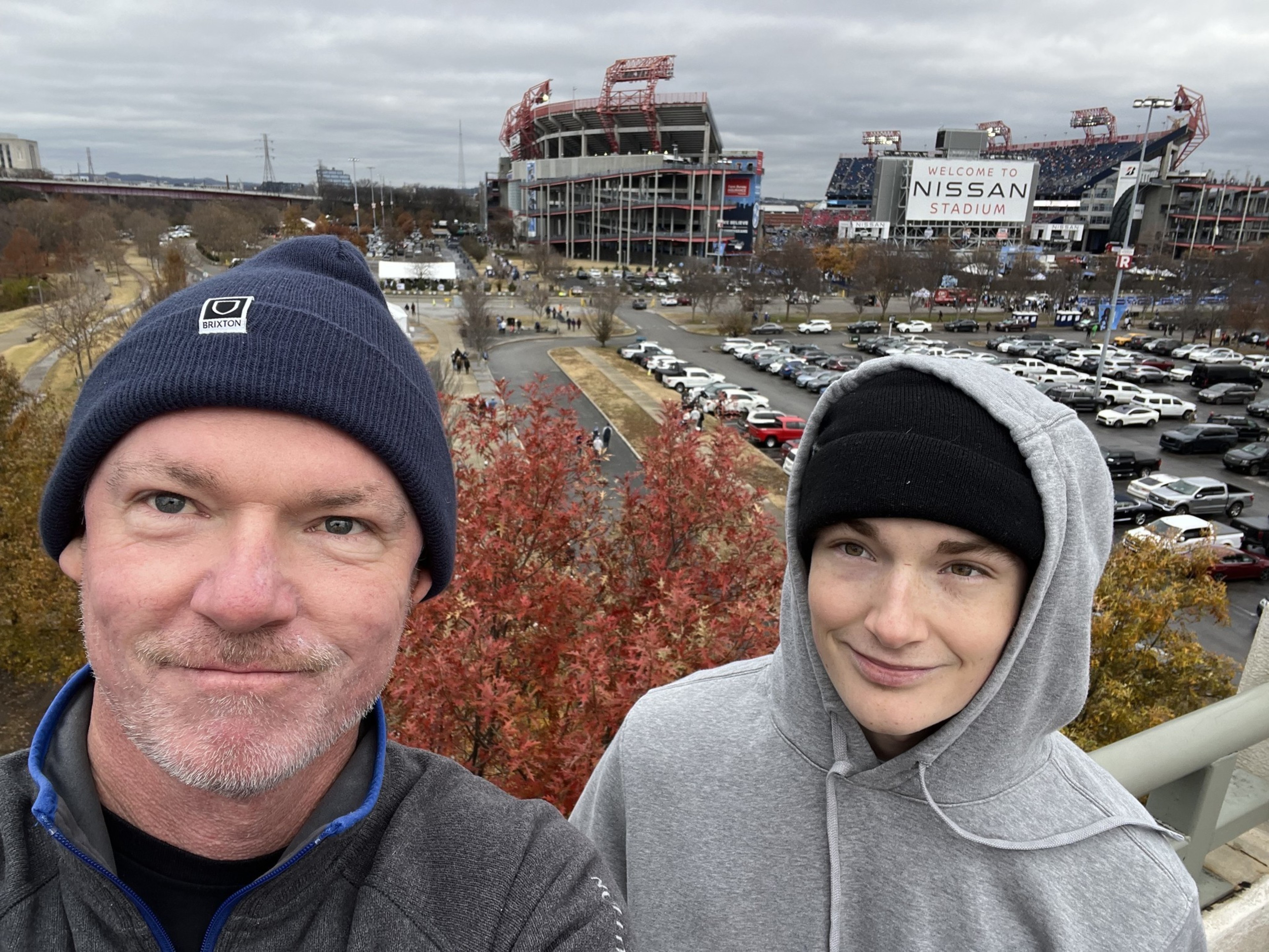 Two people wearing hats and jackets stand outdoors near colorful trees, with a parking lot and a large stadium labeled "Welcome to Nissan Stadium" in the background.