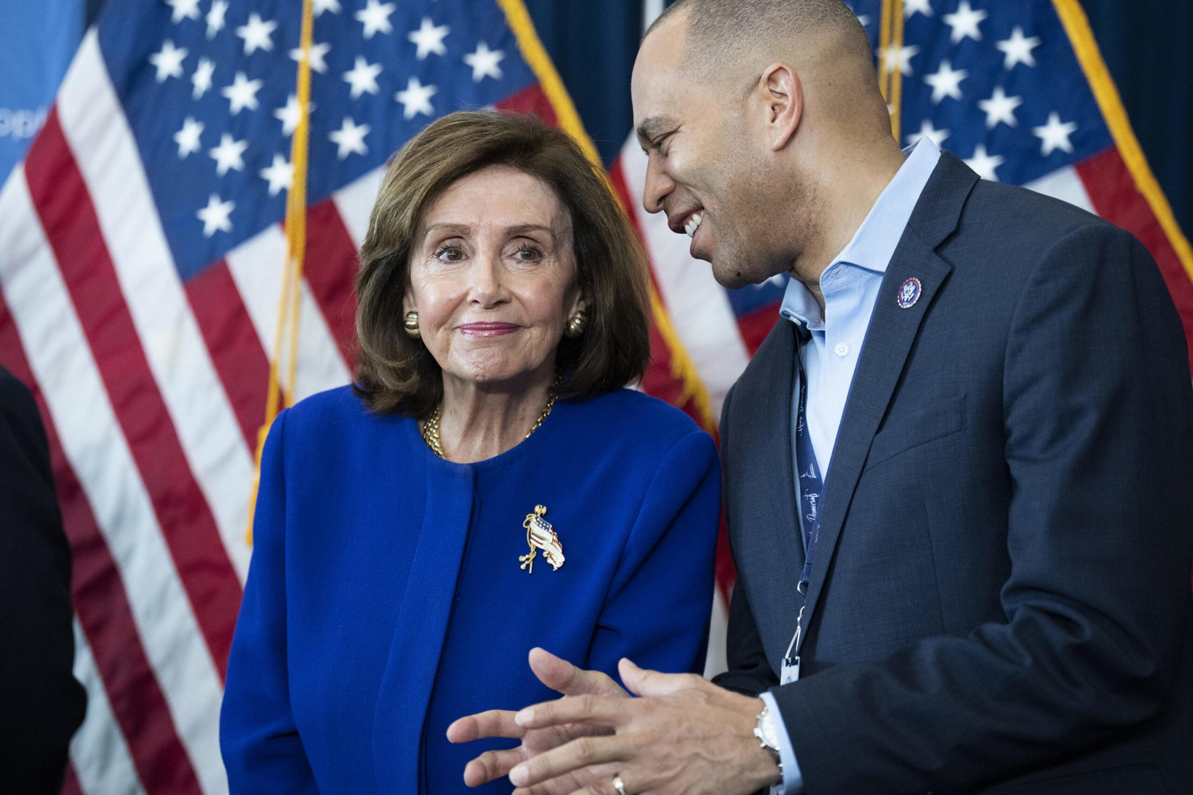 A woman in a blue suit with a brooch stands next to a man in a suit, talking and smiling. They are in front of several American flags.