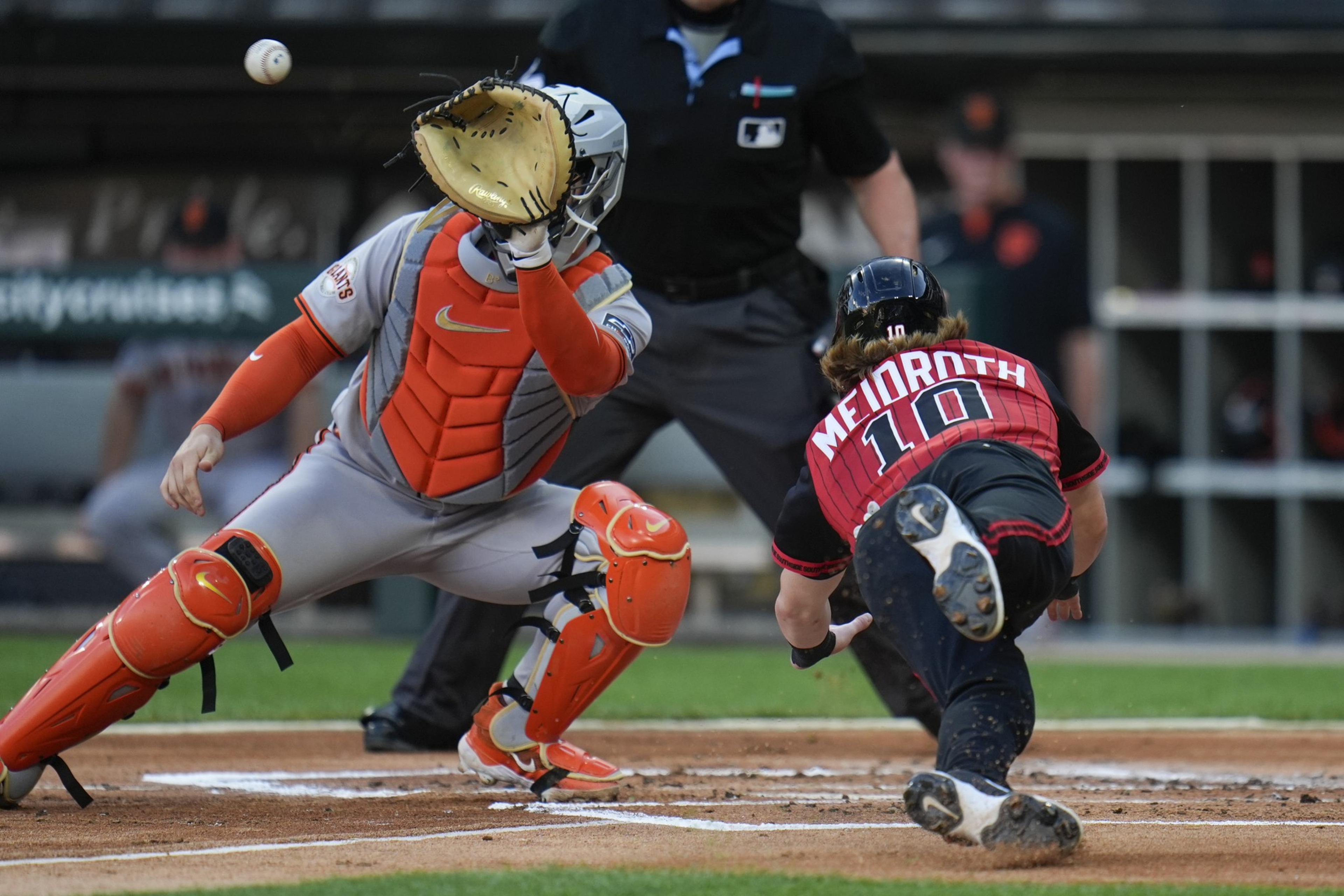 A baseball catcher in orange gear is reaching for the ball as it flies towards him. A player with &quot;MEIDROTH 10&quot; on his jersey slides into home plate.