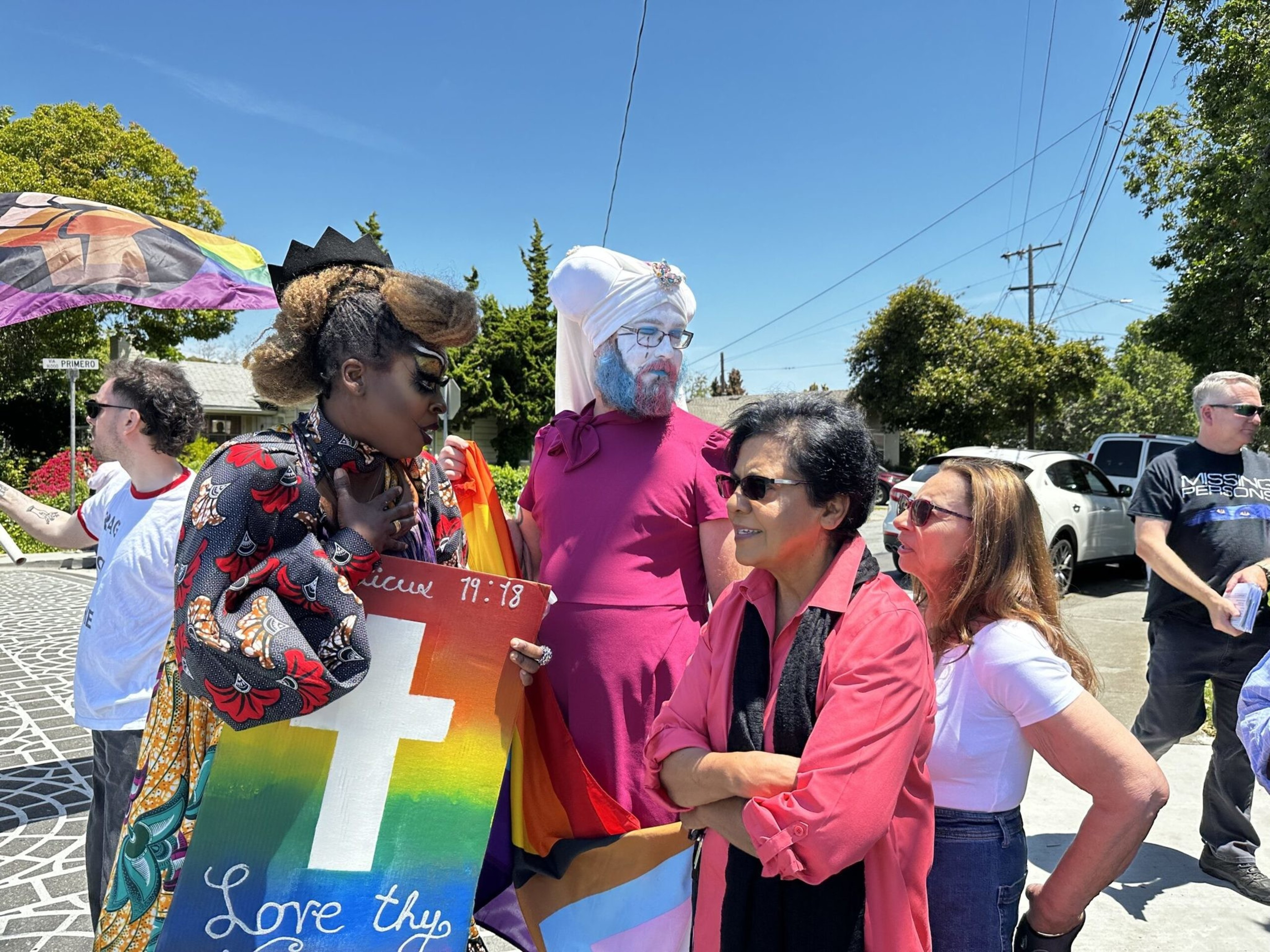 A diverse group of people stands outdoors, including a person in colorful attire holding a rainbow cross sign with "Love thy neighbor" written on it.