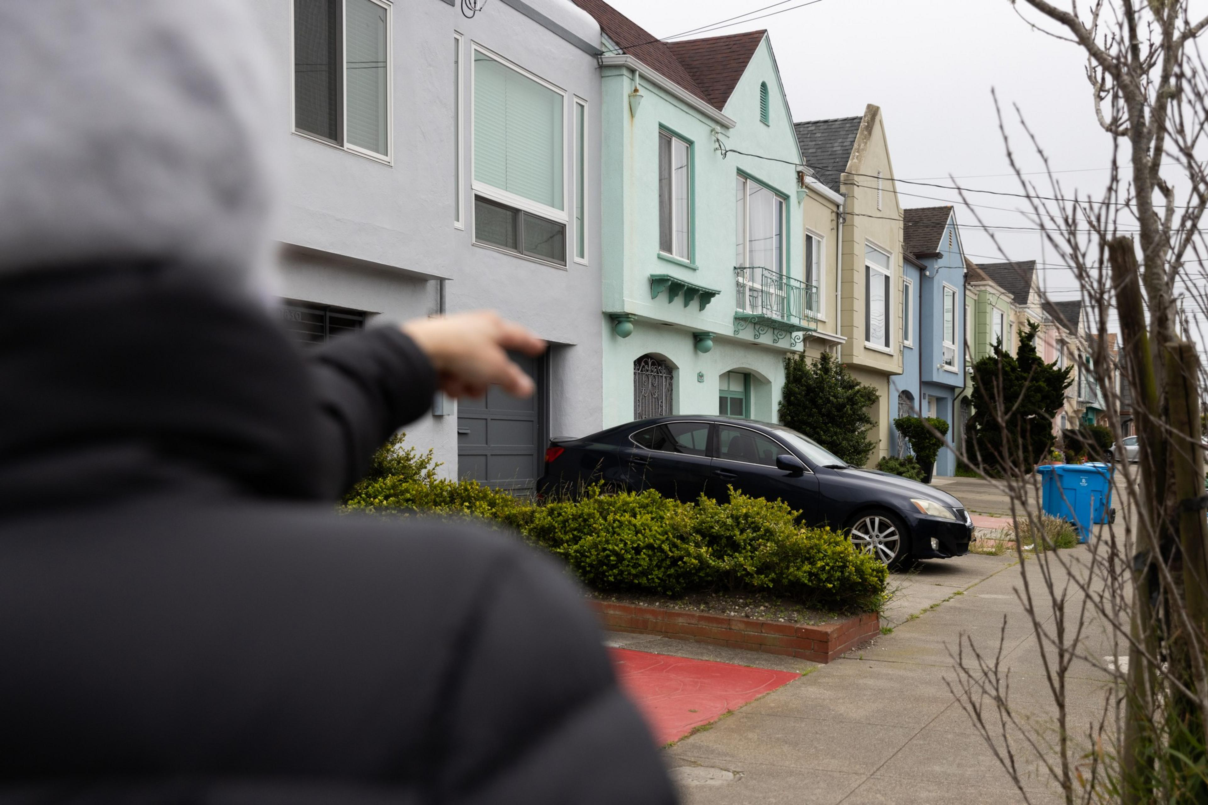 A person's pointing hand, colorful houses, a parked car, and a tree-lined sidewalk.