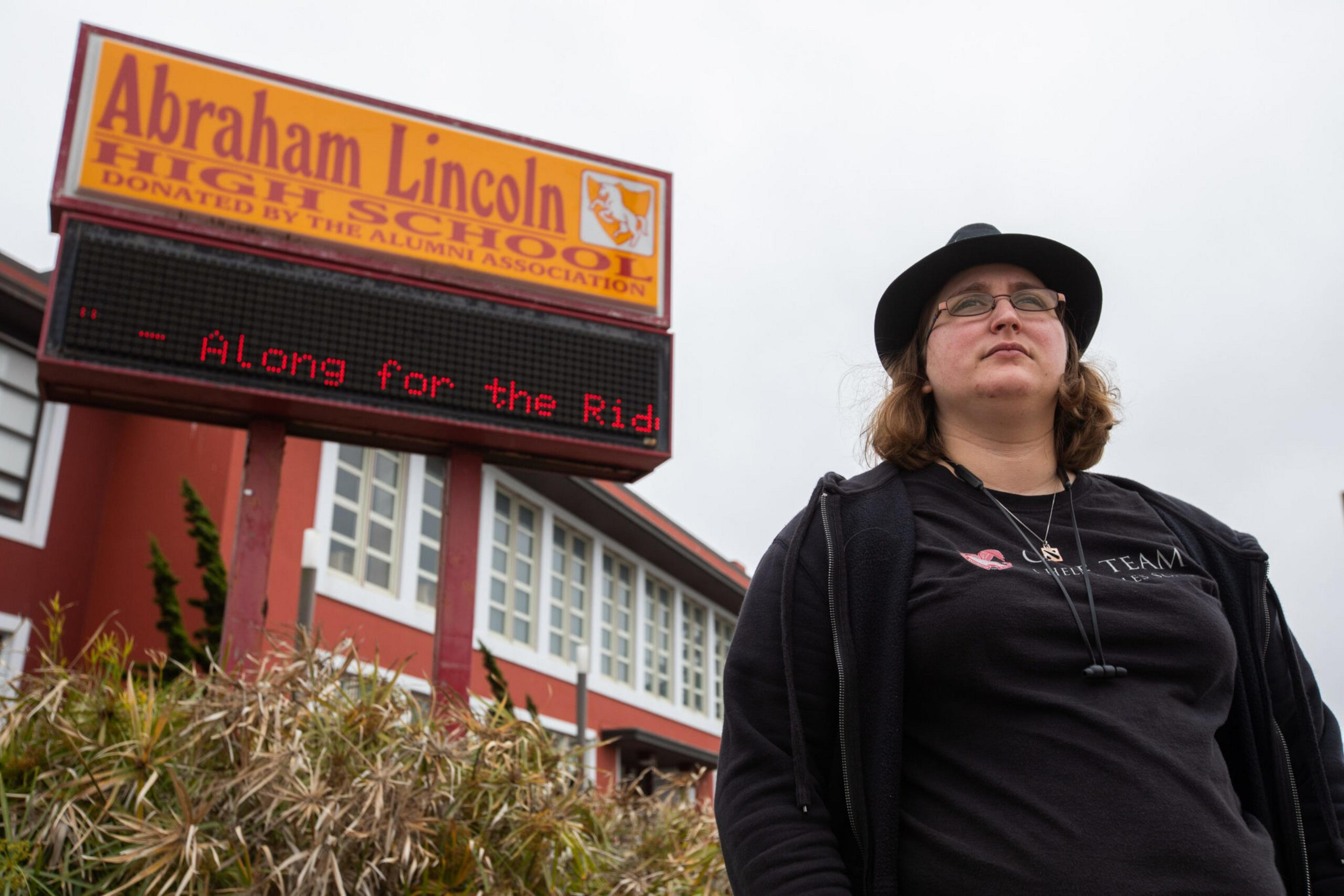 A person wearing glasses and a black hat stands in front of Abraham Lincoln High School with a digital message board partially showing text.