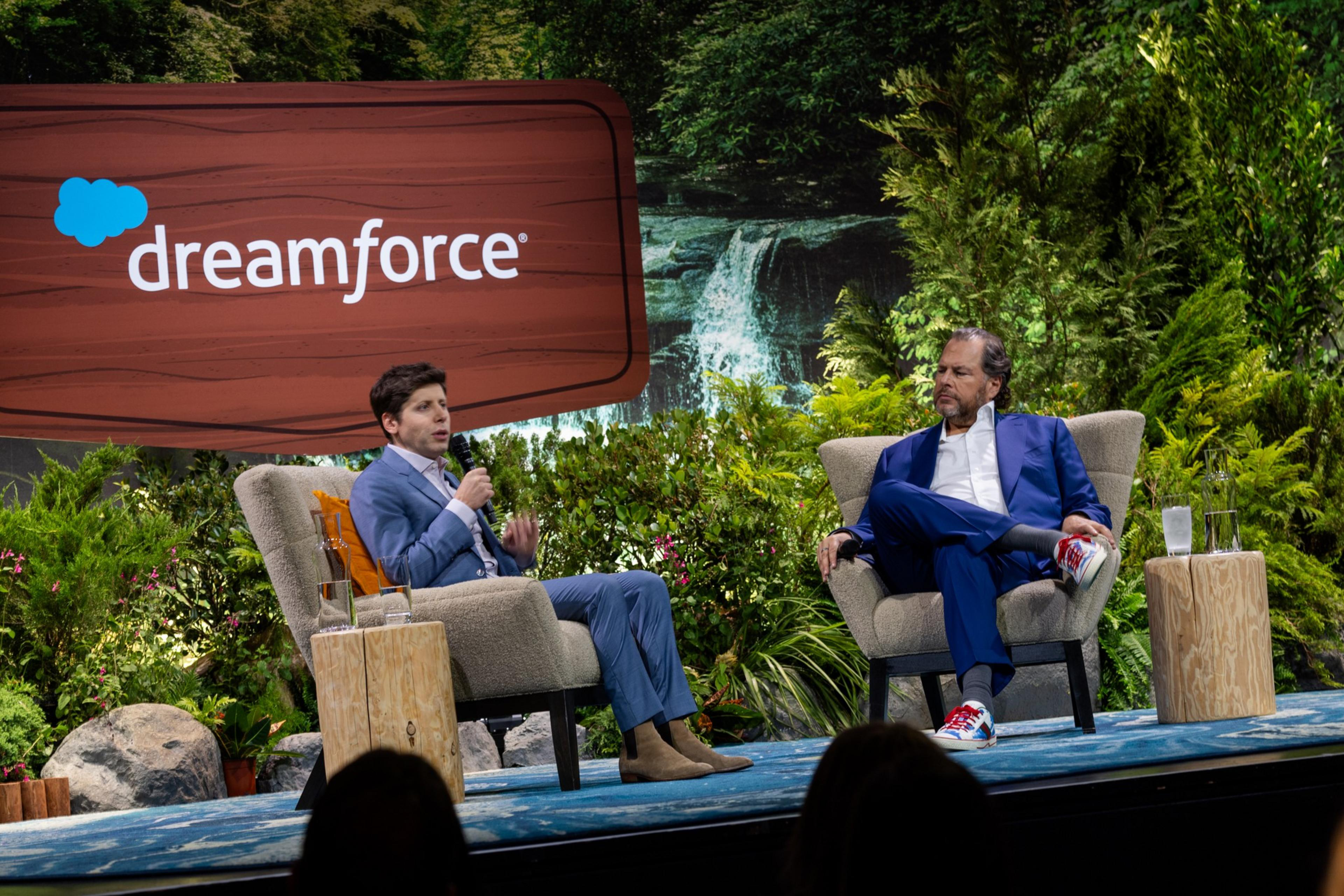 Two men in blue suits sit on armchairs on a stage with a forest and waterfall backdrop, under a large "dreamforce" sign.