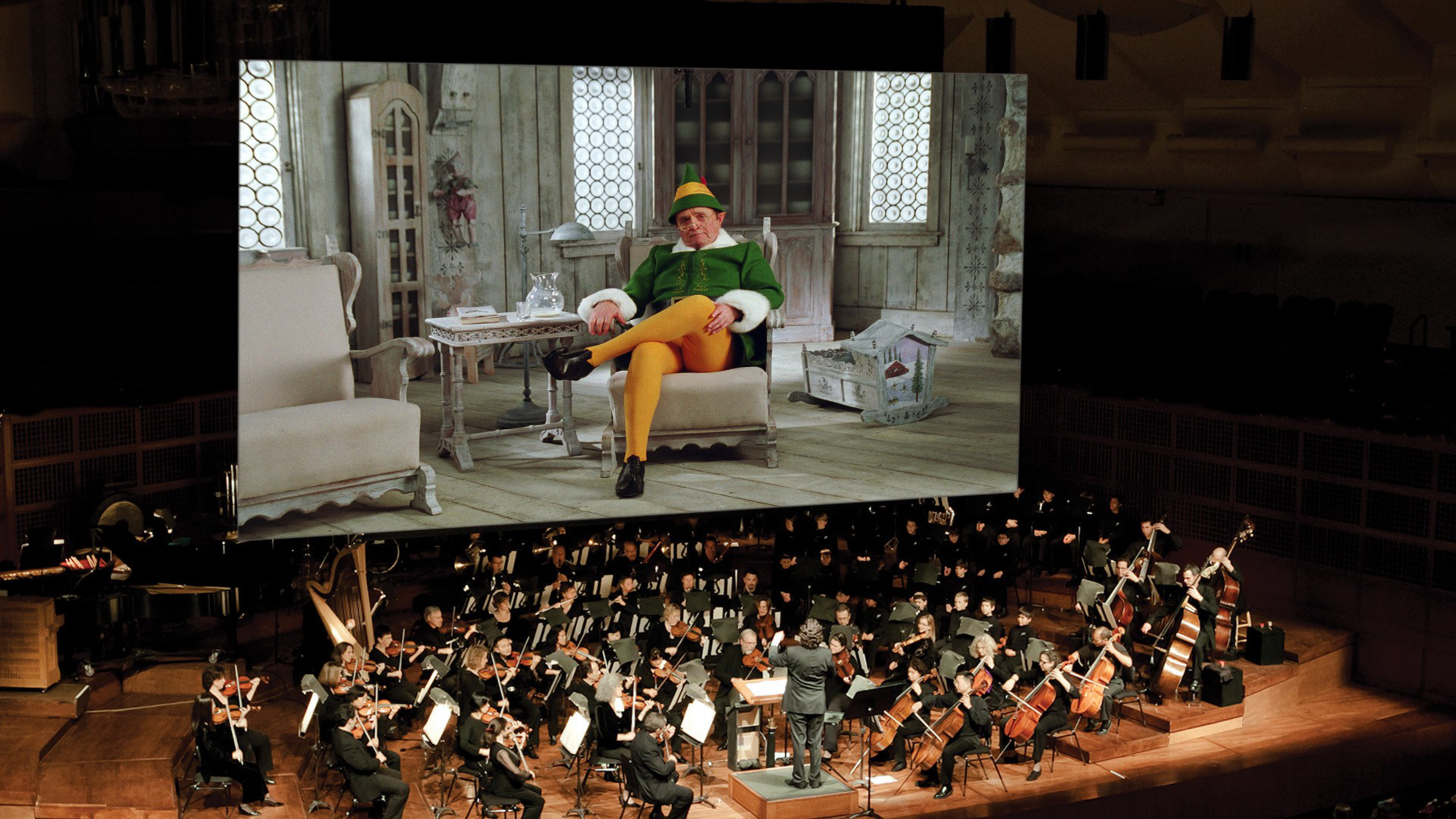 An orchestra plays on a wooden stage while a large screen above shows a man in a green and yellow costume sitting with crossed legs in a vintage room.